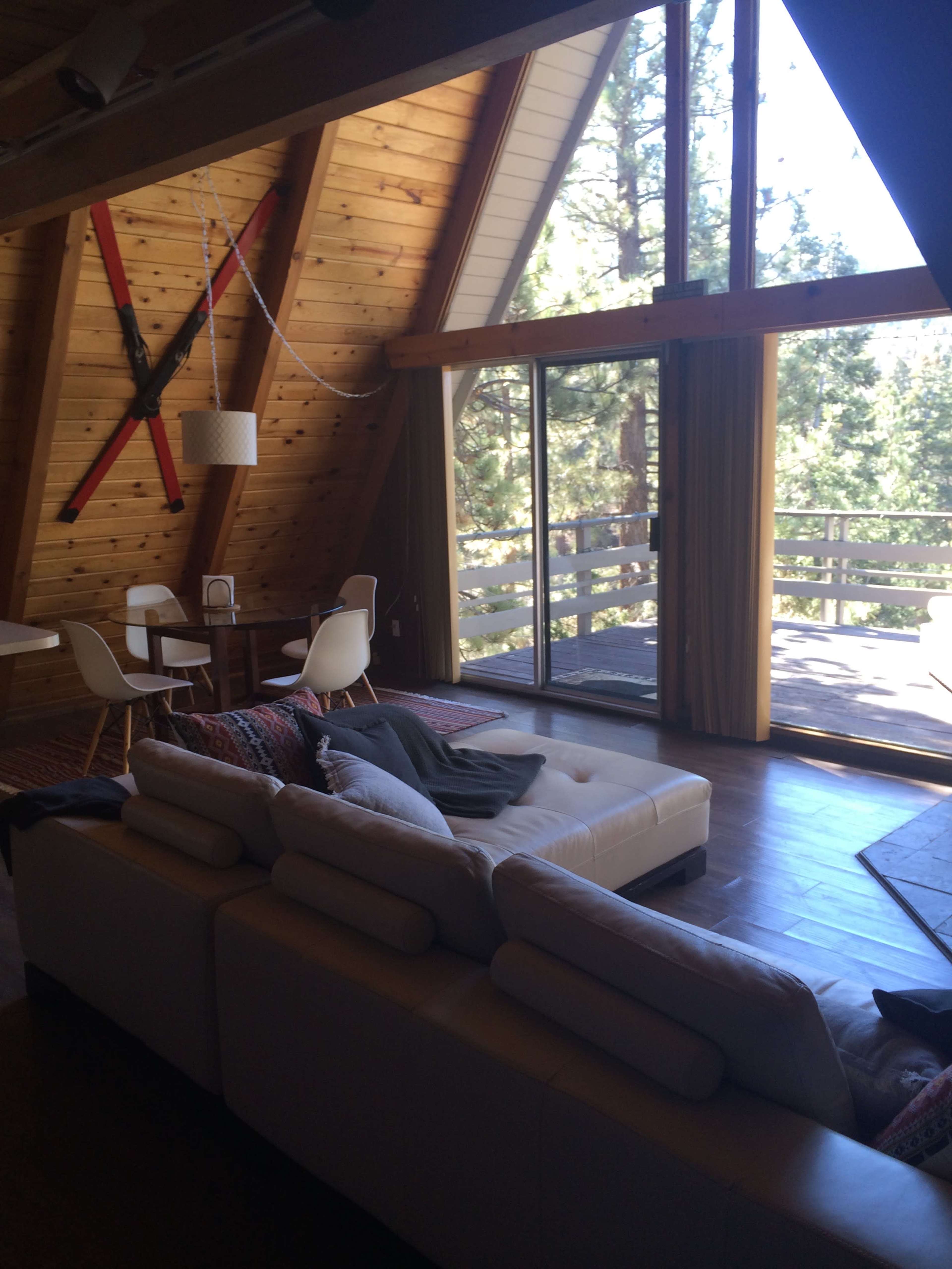 The image shows a modern living room in a triangular-shaped cabin with wooden walls, large windows, and a white sectional sofa facing a sliding glass door that opens to a balcony.
