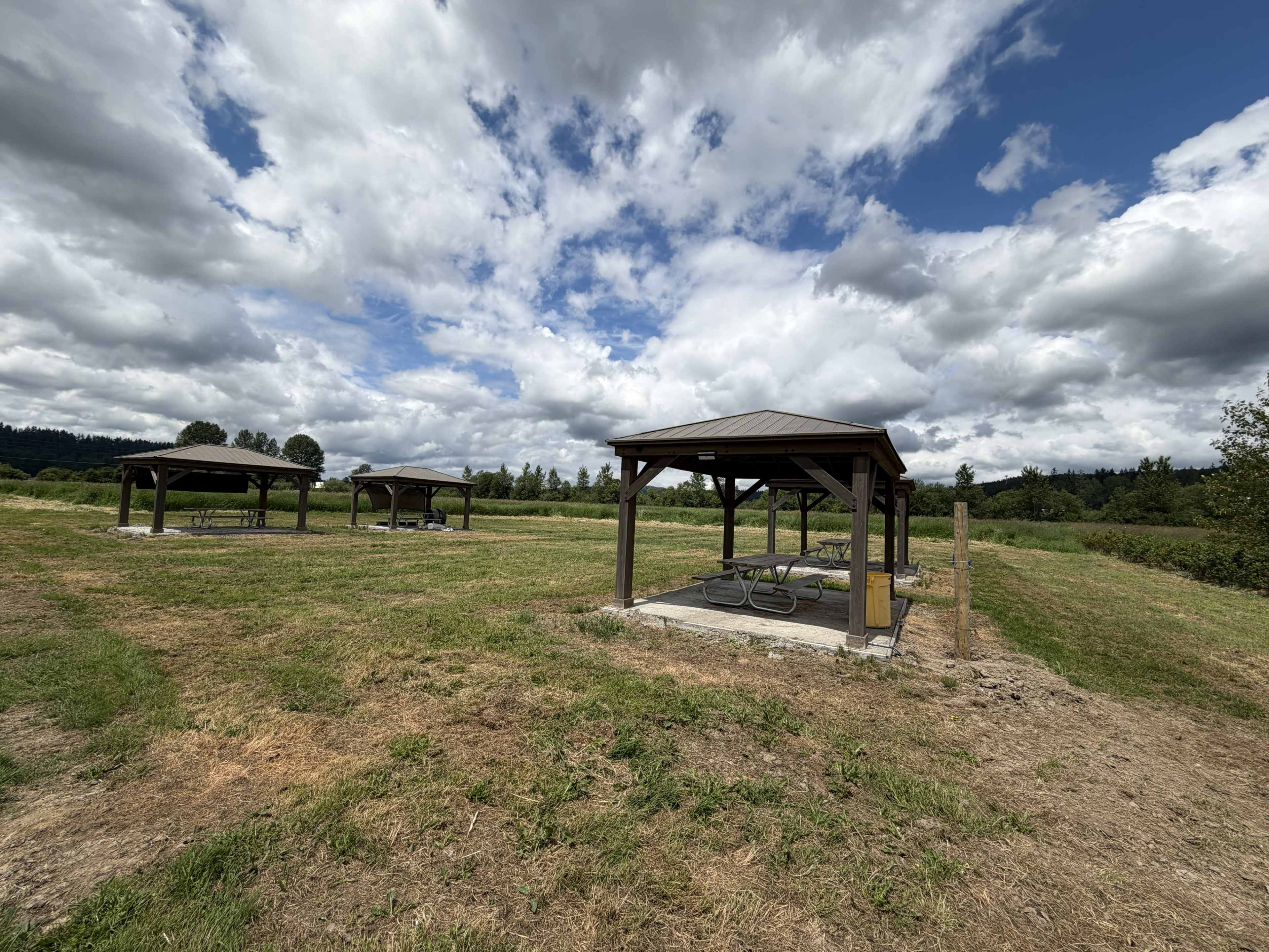 The image shows several wooden picnic shelters on a grassy field under a partly cloudy sky.