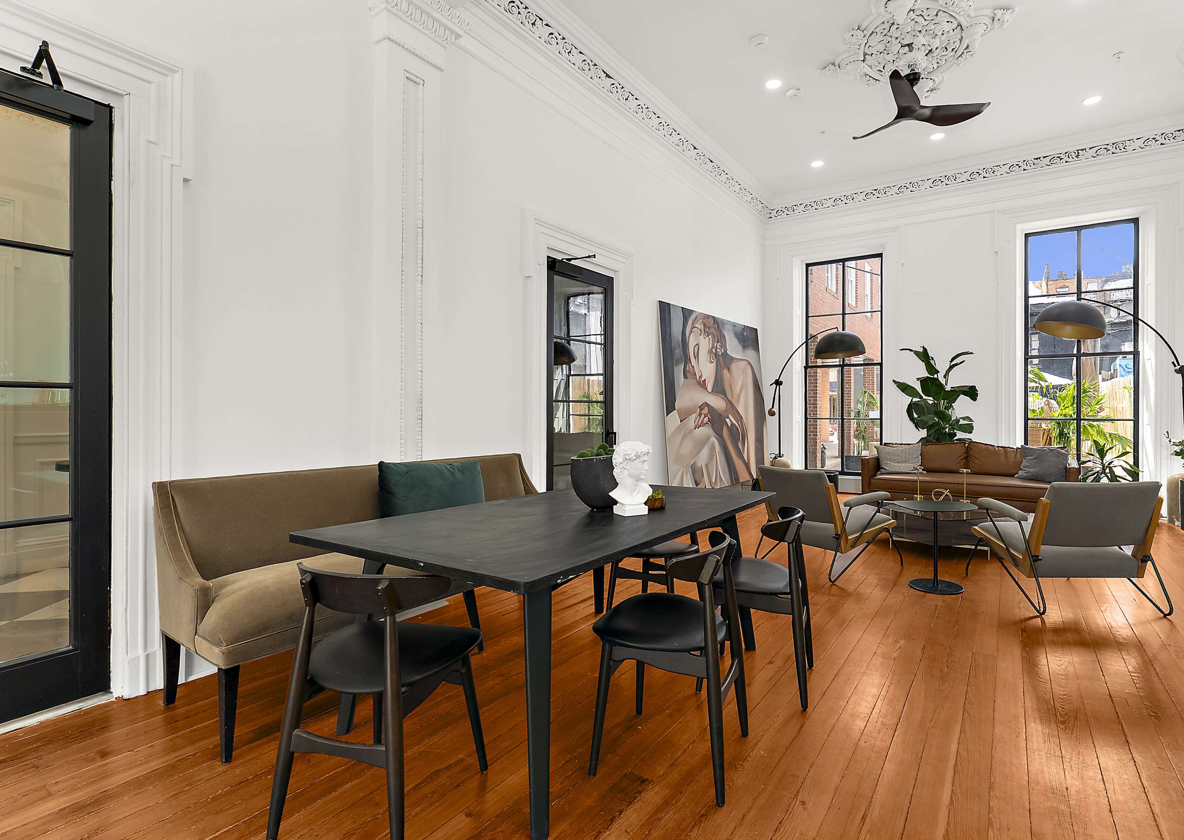 The image shows a modern living space with a dining table, black chairs, large windows, a couch, artwork on the wall, and decorative plants.