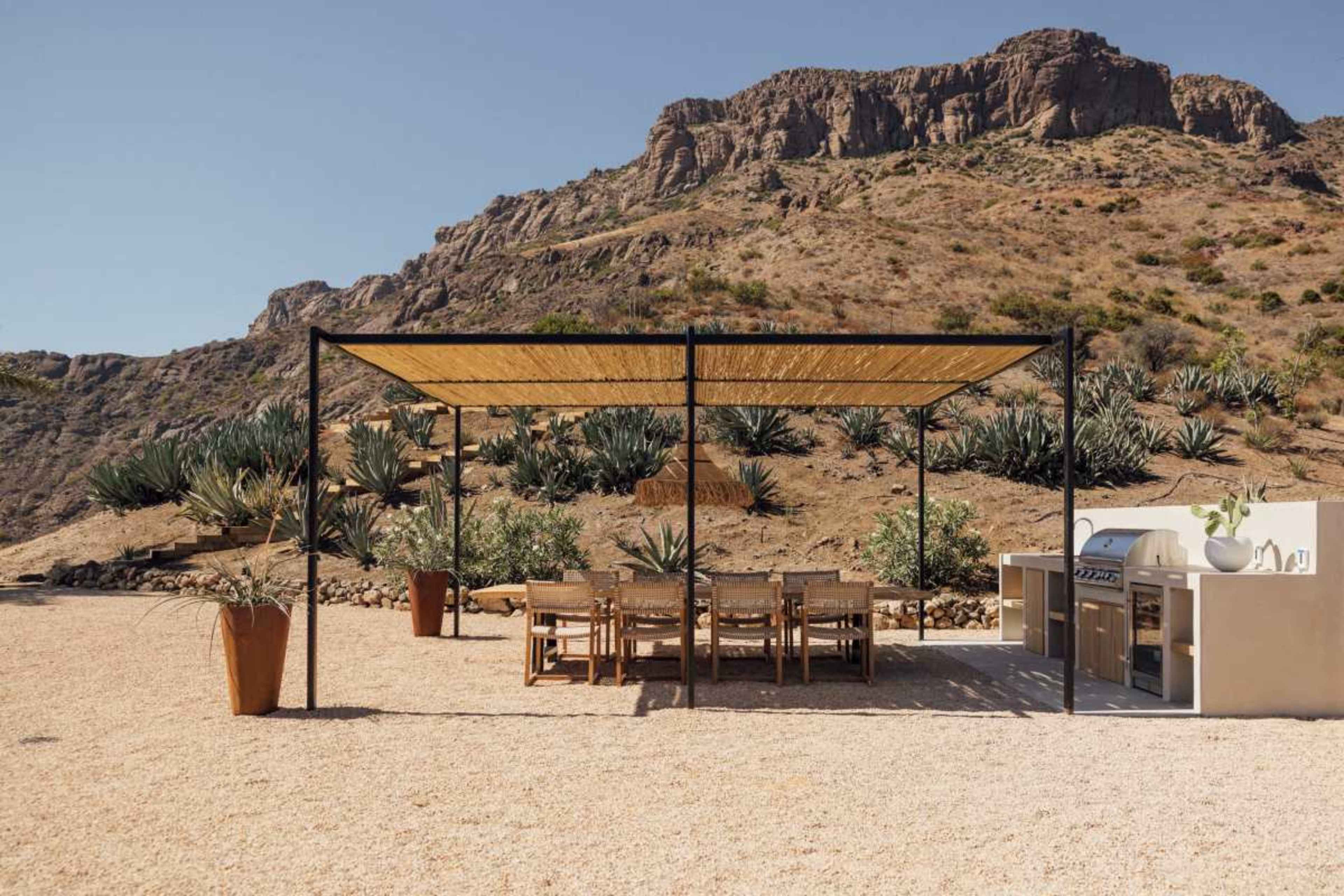 A covered dining area with a large table and chairs stands in front of a rocky hillside, surrounded by desert vegetation.