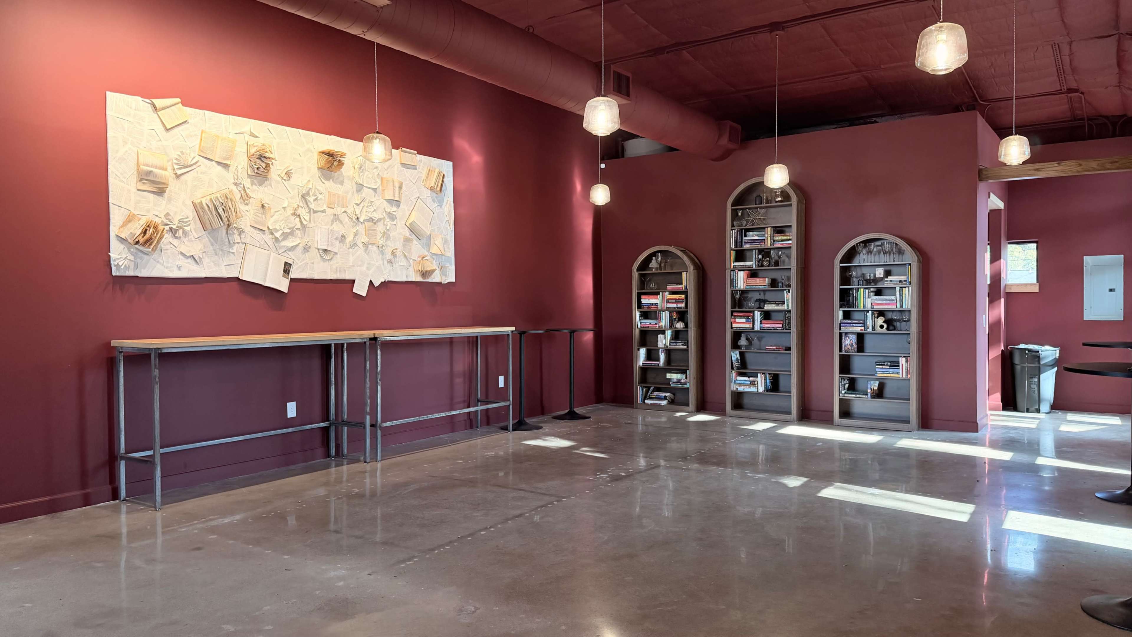 The image shows a room with a deep red wall, featuring a textured artwork on one side and bookshelves with books on the other, along with a polished concrete floor and pendant lighting.
