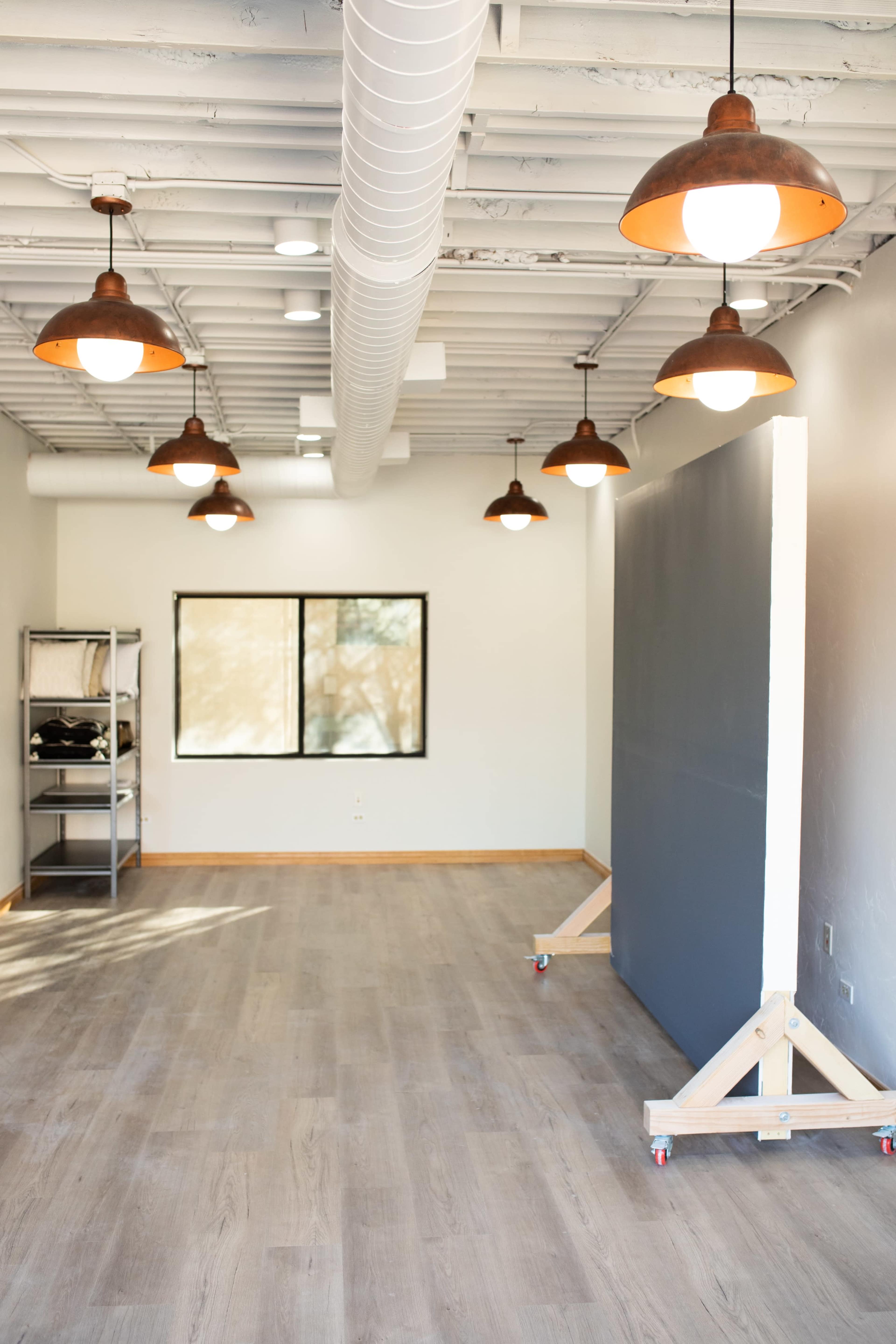 A spacious room with wooden flooring, featuring overhead industrial-style pendant lights and a large, movable gray panel on a wheeled stand.