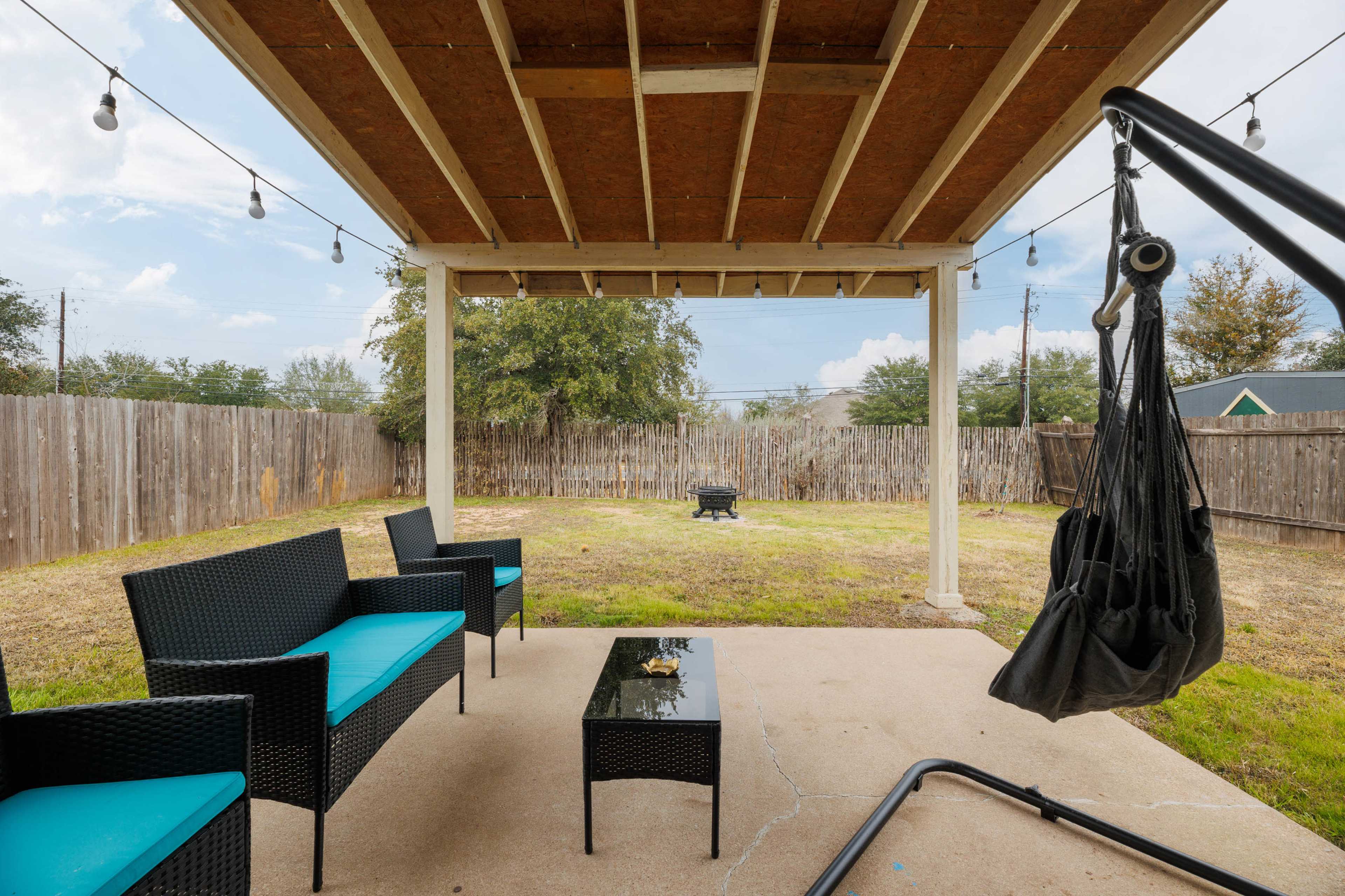 A covered patio area features black wicker furniture with teal cushions, a glass-top coffee table, and a hanging chair, overlooking a spacious grassy yard enclosed by a wooden fence.