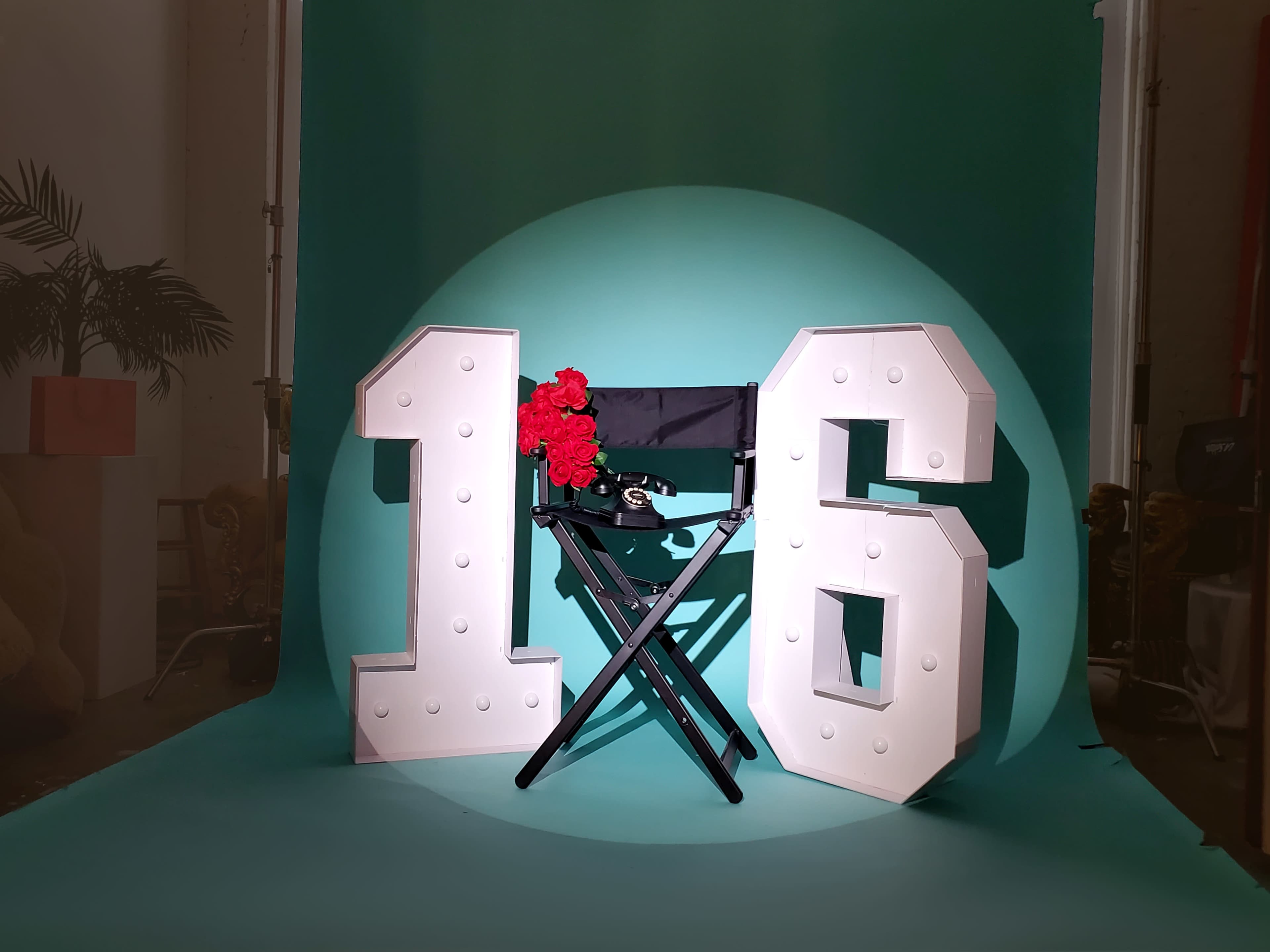 The image features large, illuminated numbers "1" and "6" positioned beside a black director's chair adorned with red roses, set against a teal backdrop.