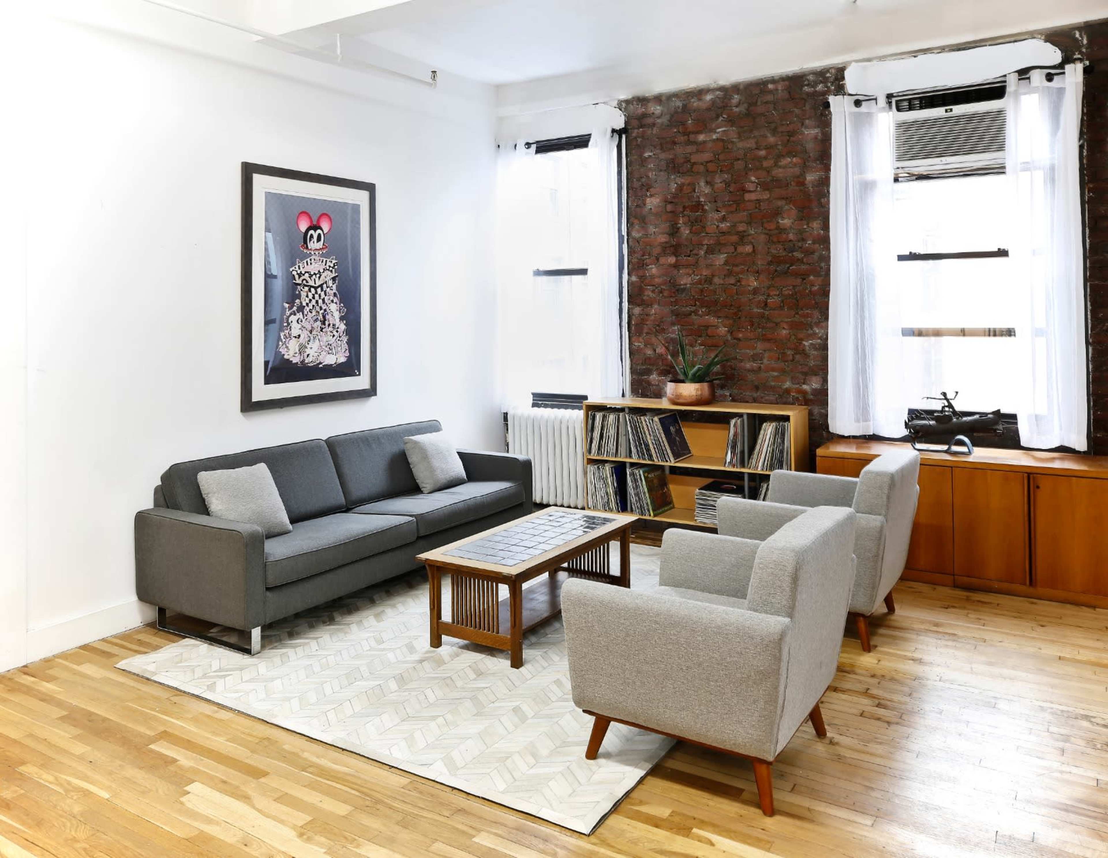 A modern living room featuring a grey sofa, two light grey armchairs, a wooden coffee table, a bookshelf filled with records, and exposed brick walls.