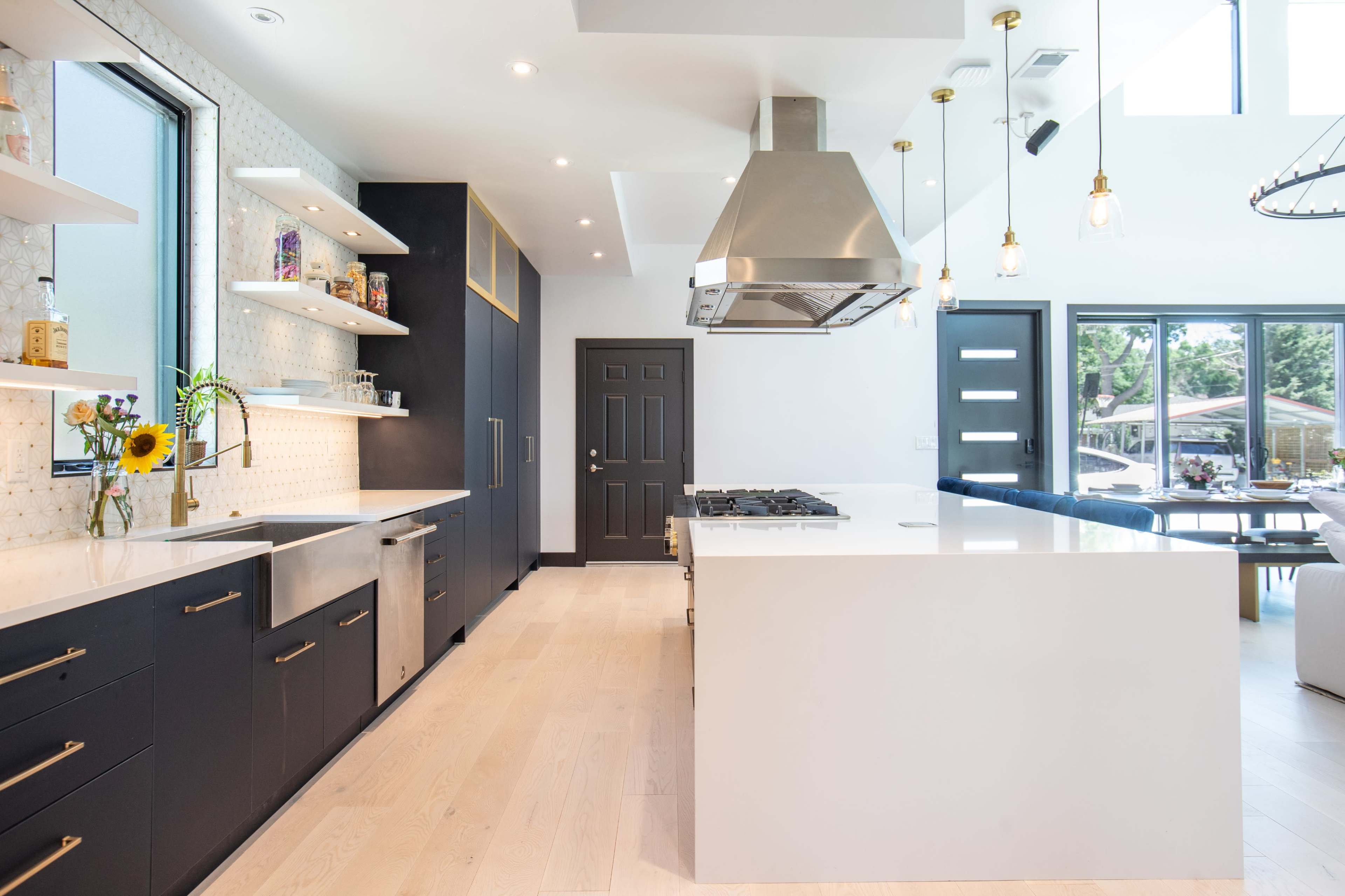 A modern kitchen features white countertops, dark cabinets, a central island with a stovetop, and large windows allowing natural light.