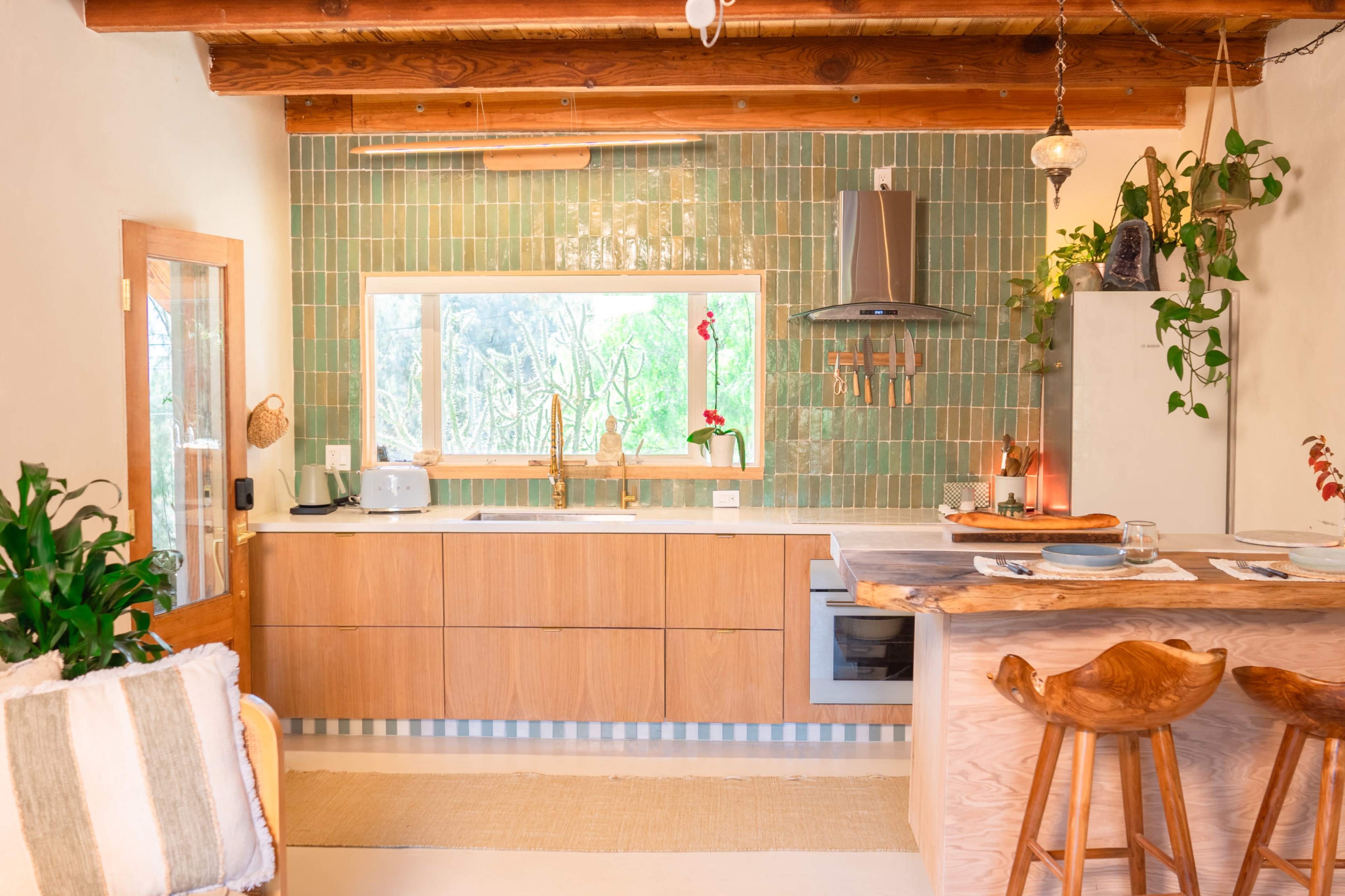 The image shows a modern kitchen featuring wooden cabinetry, a stone countertop, a large window with greenery visible outside, and decorative tiles on the wall.