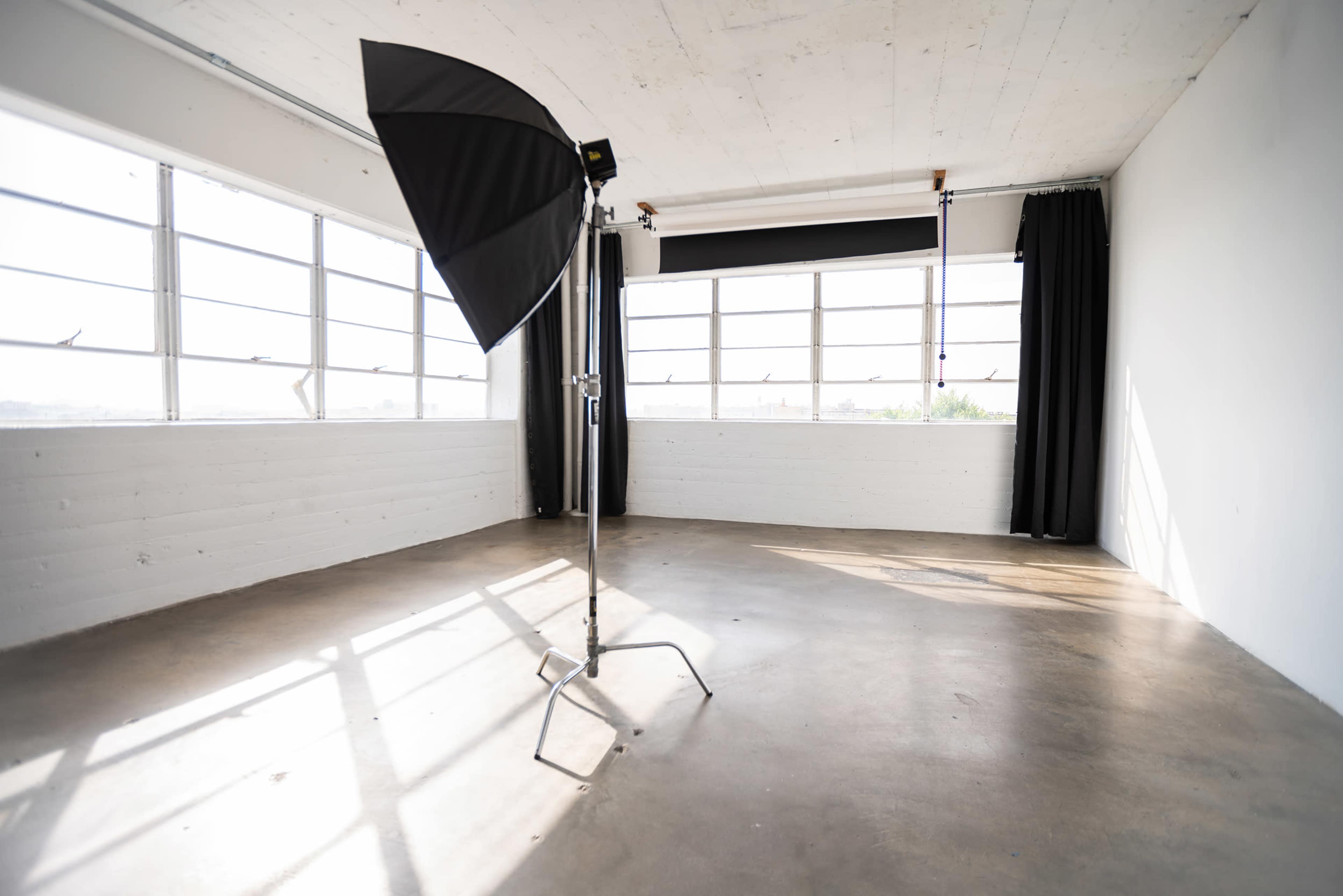 A spacious, empty studio with a large window and a black umbrella light on a stand.
