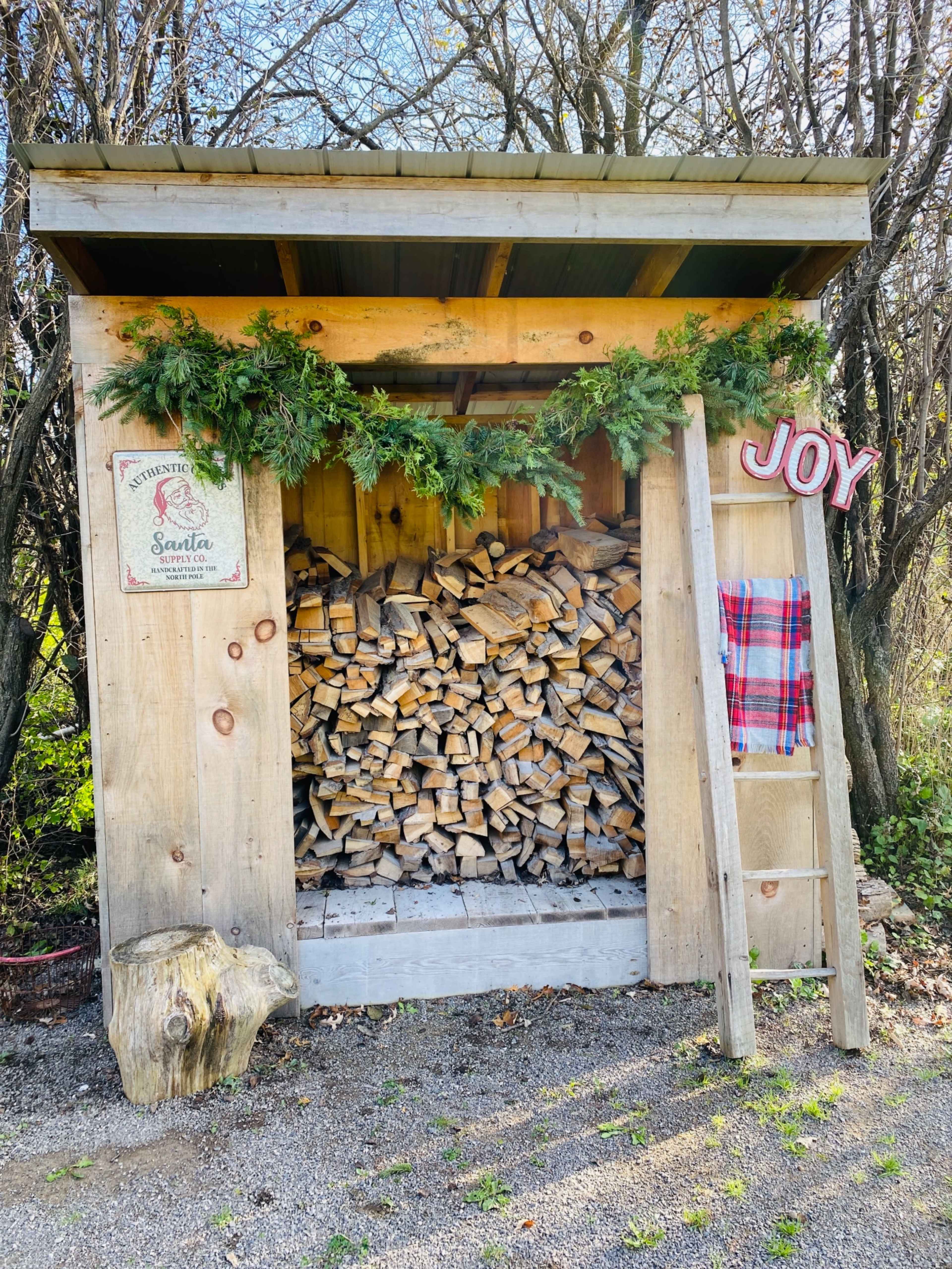 A wooden shed filled with neatly stacked firewood and adorned with greenery, a festive sign, and a ladder leaning against it.