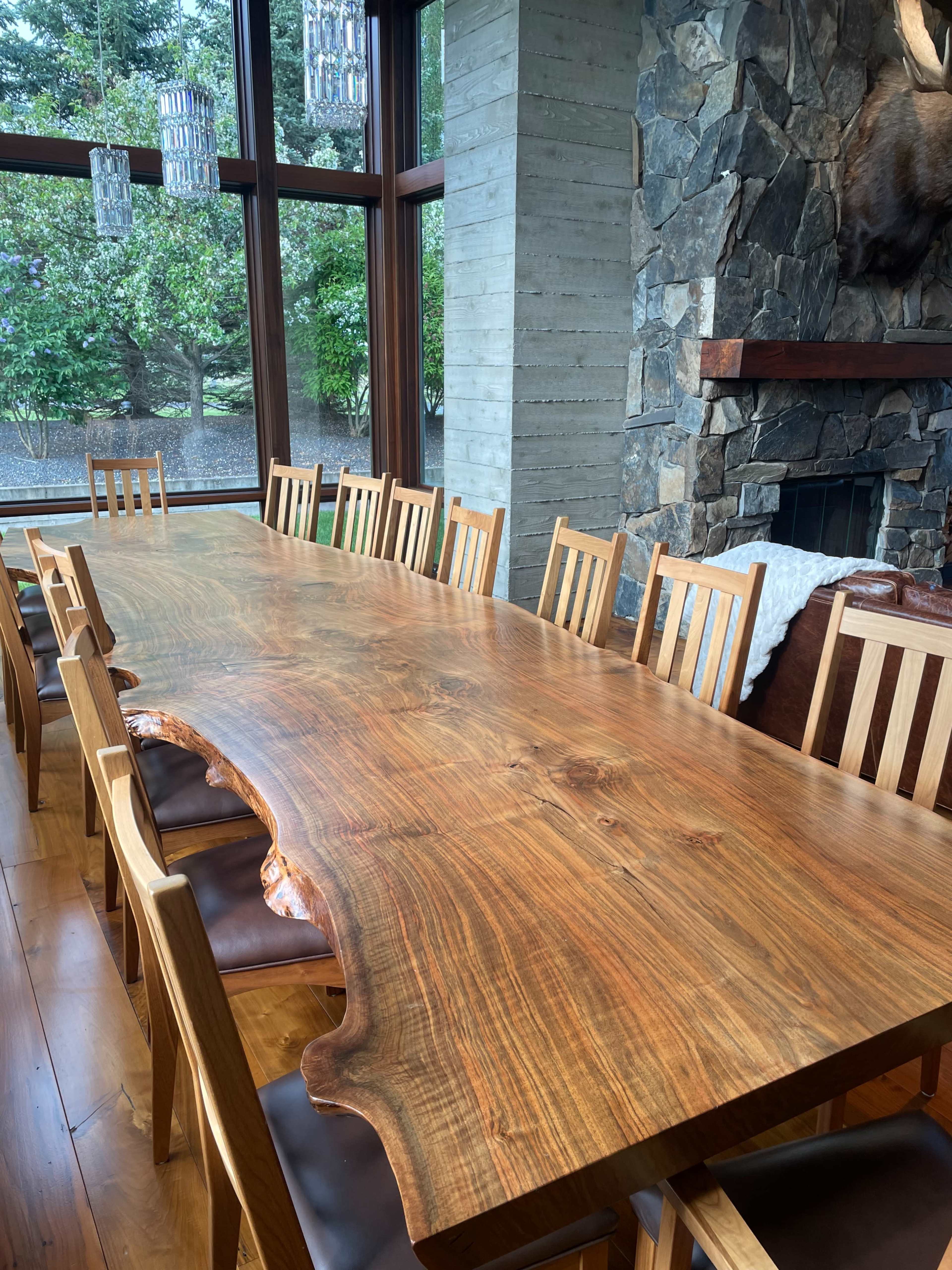 The image shows a large wooden dining table surrounded by wooden chairs in a well-lit room with windows overlooking greenery.