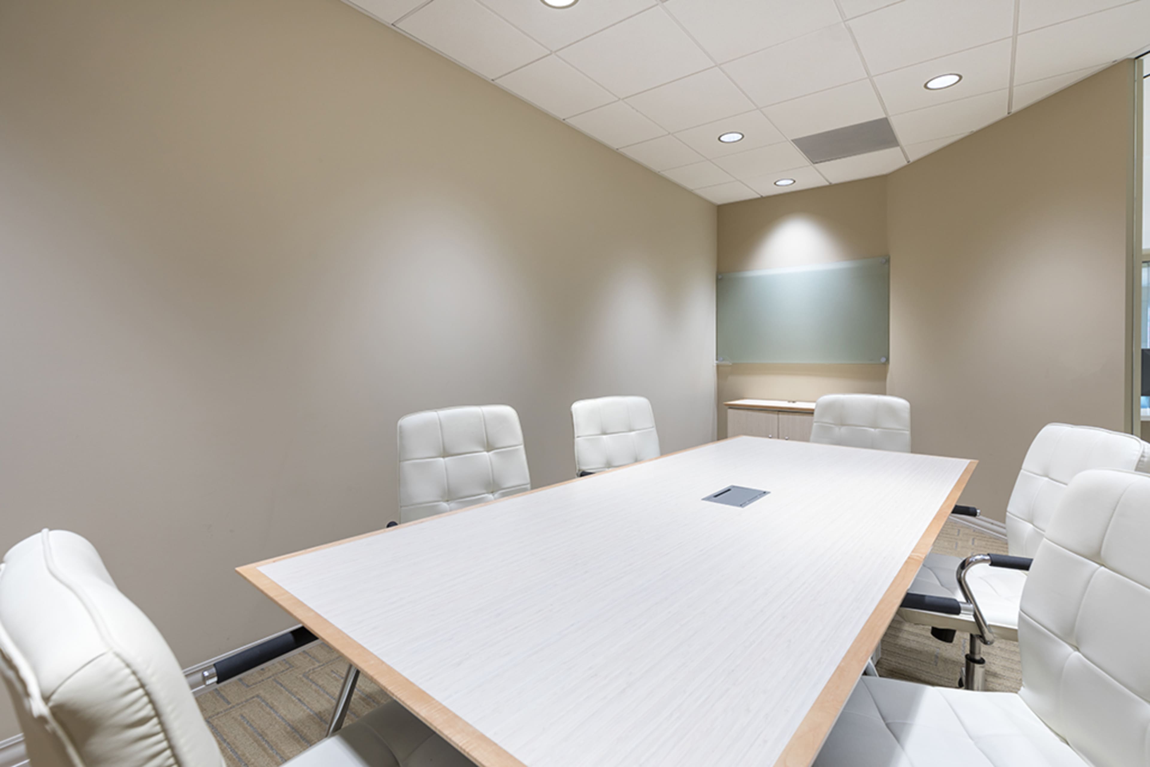 A modern conference room features a large wooden table surrounded by white leather chairs, with minimal decor on the walls.
