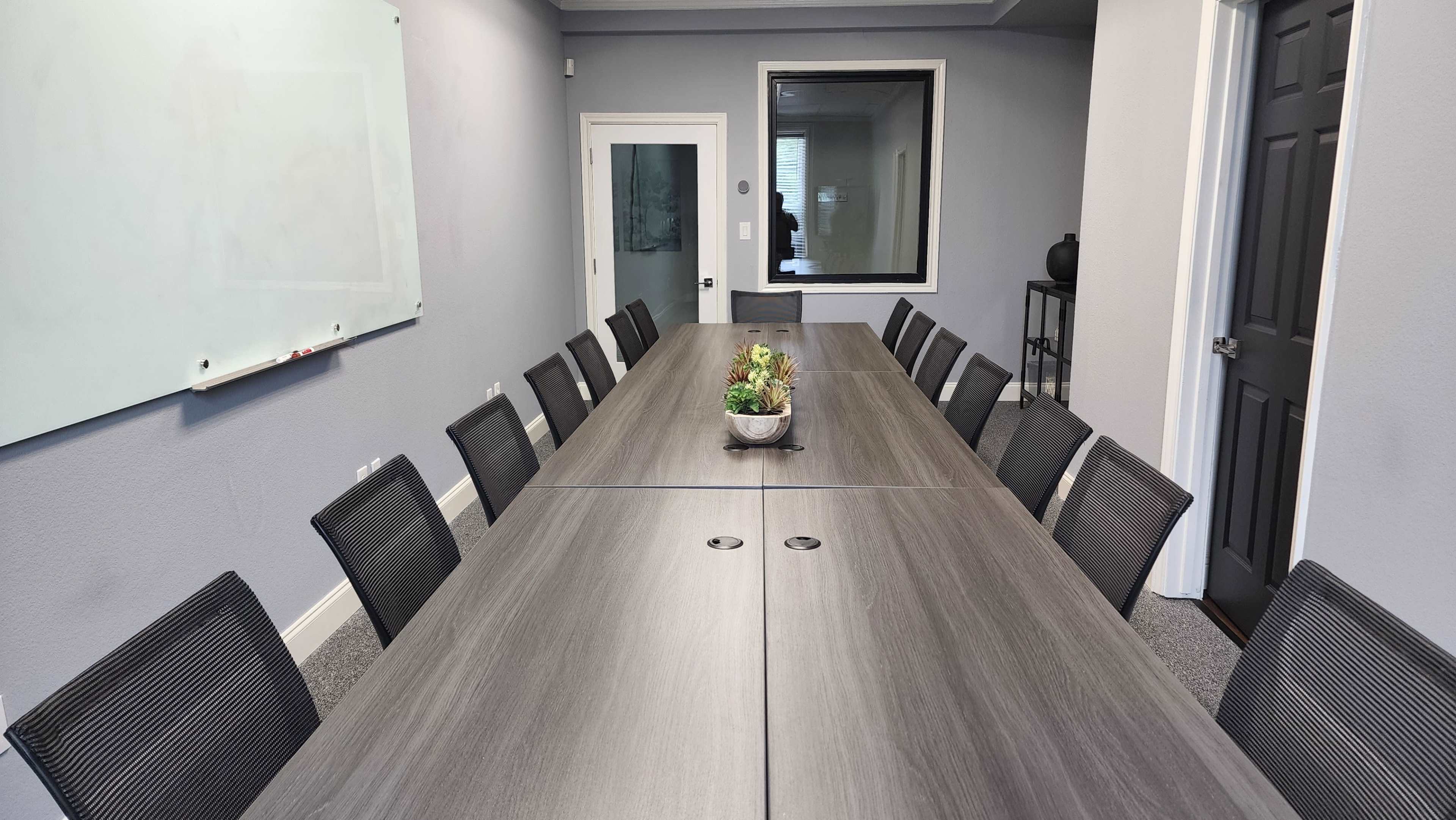 A long conference table surrounded by black mesh chairs is set up in a meeting room with gray walls and a whiteboard.