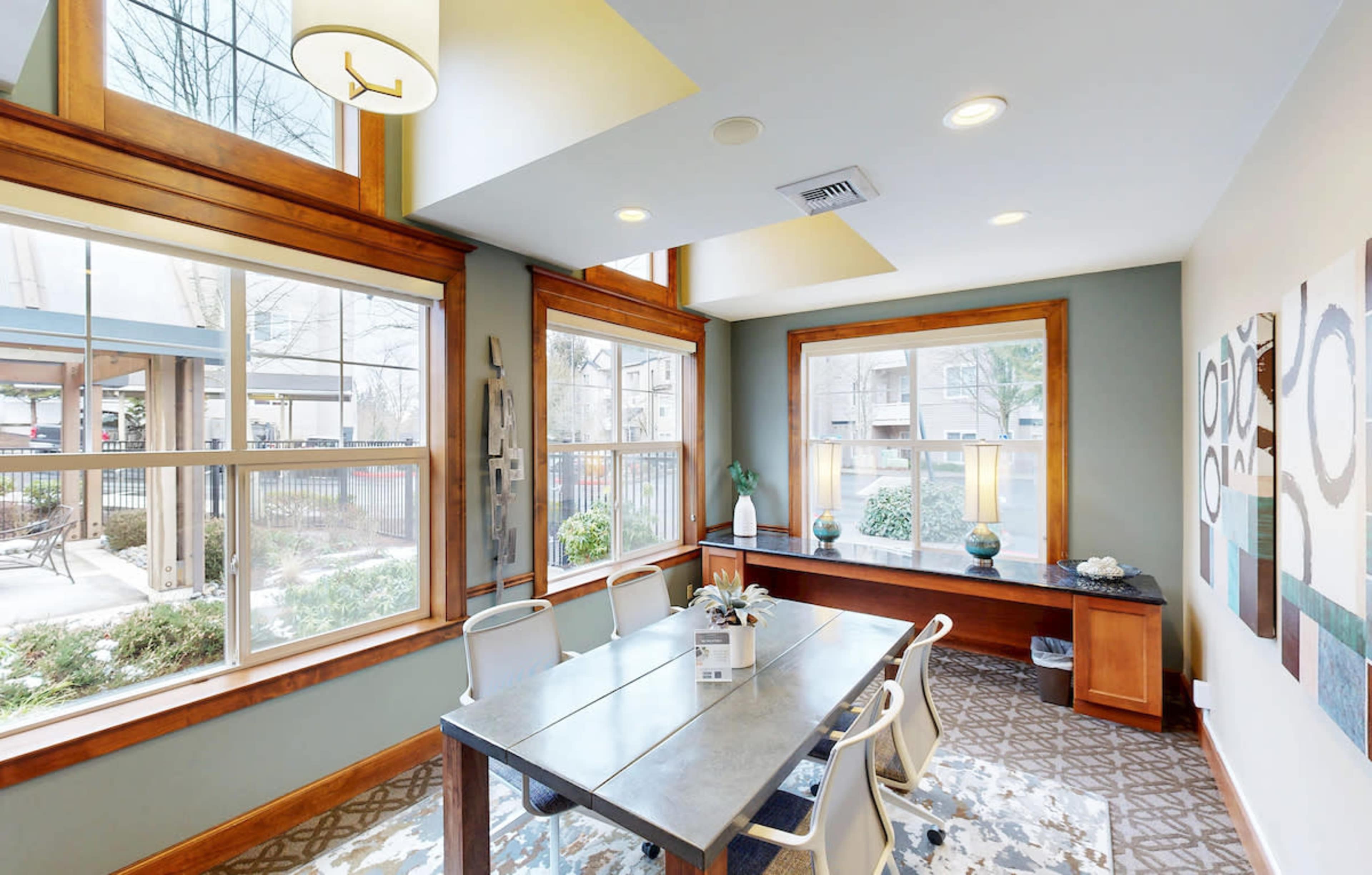 The image shows a well-lit dining area with a wooden table and chairs, surrounded by large windows overlooking a landscaped outdoor space.