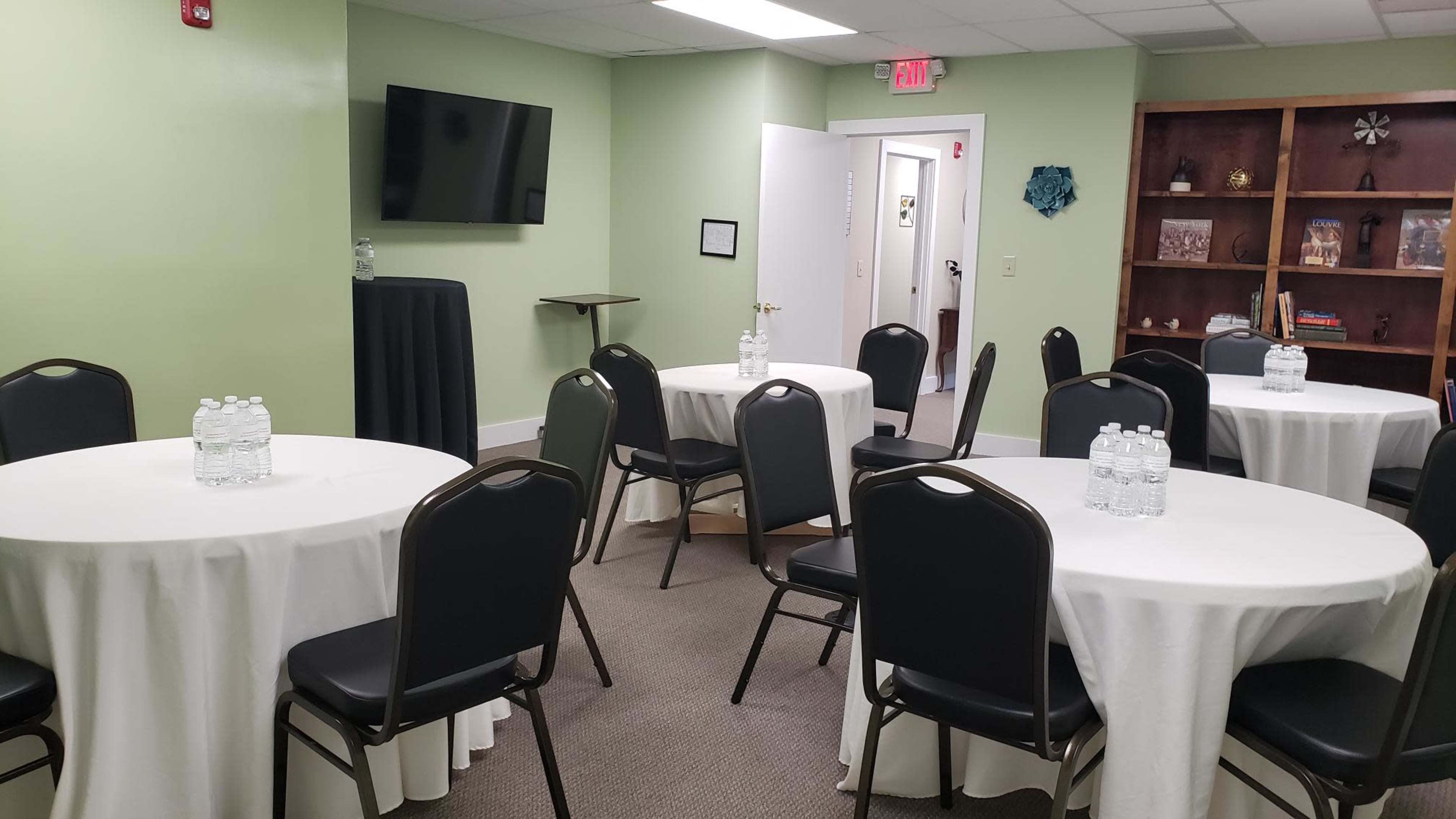 The room features several round tables covered with white tablecloths, each adorned with bottles of water, and a black table in the corner, complemented by a television on the wall.