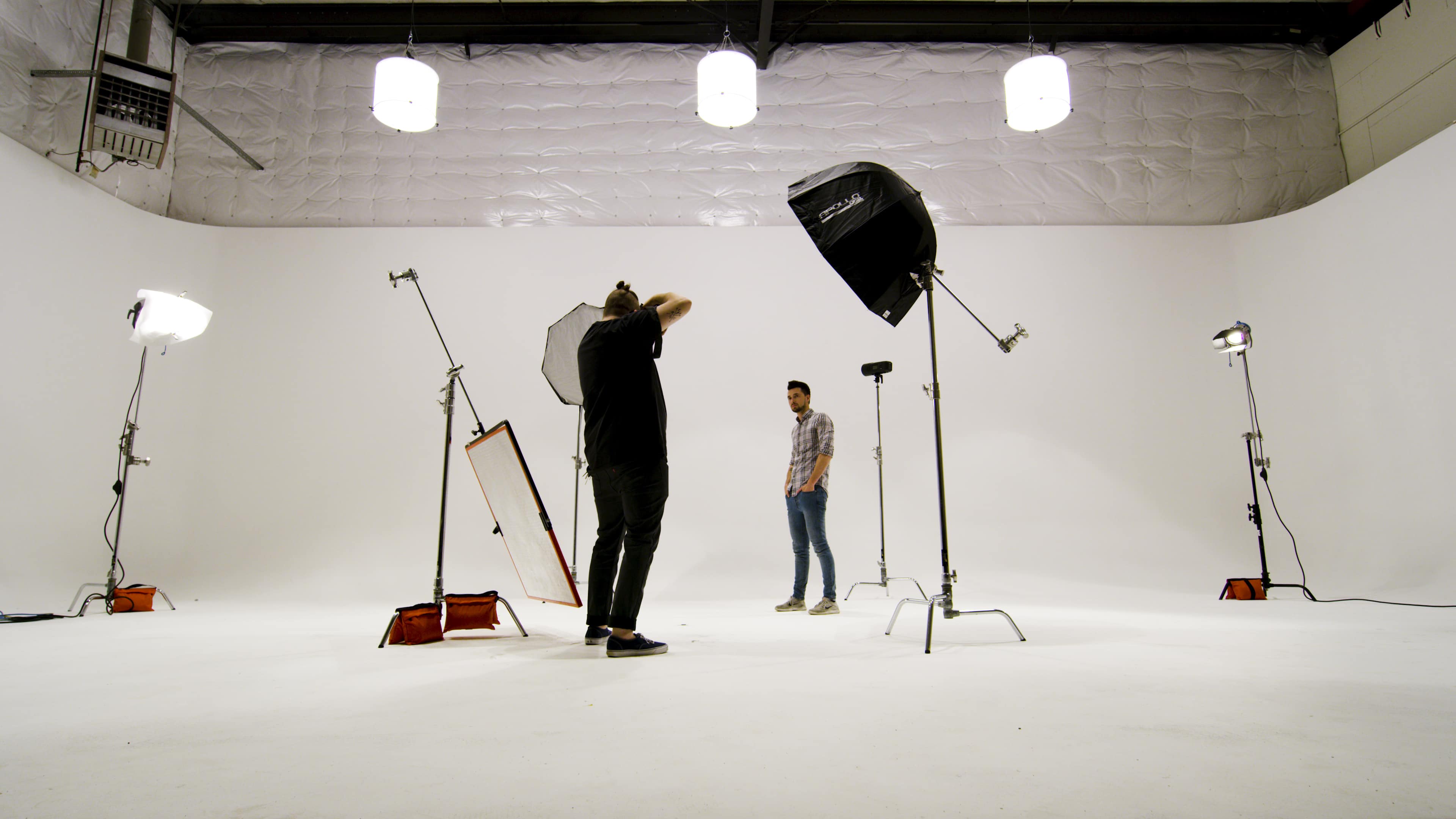 A photographer is adjusting equipment in a spacious, well-lit studio while a model stands in front of a white backdrop.