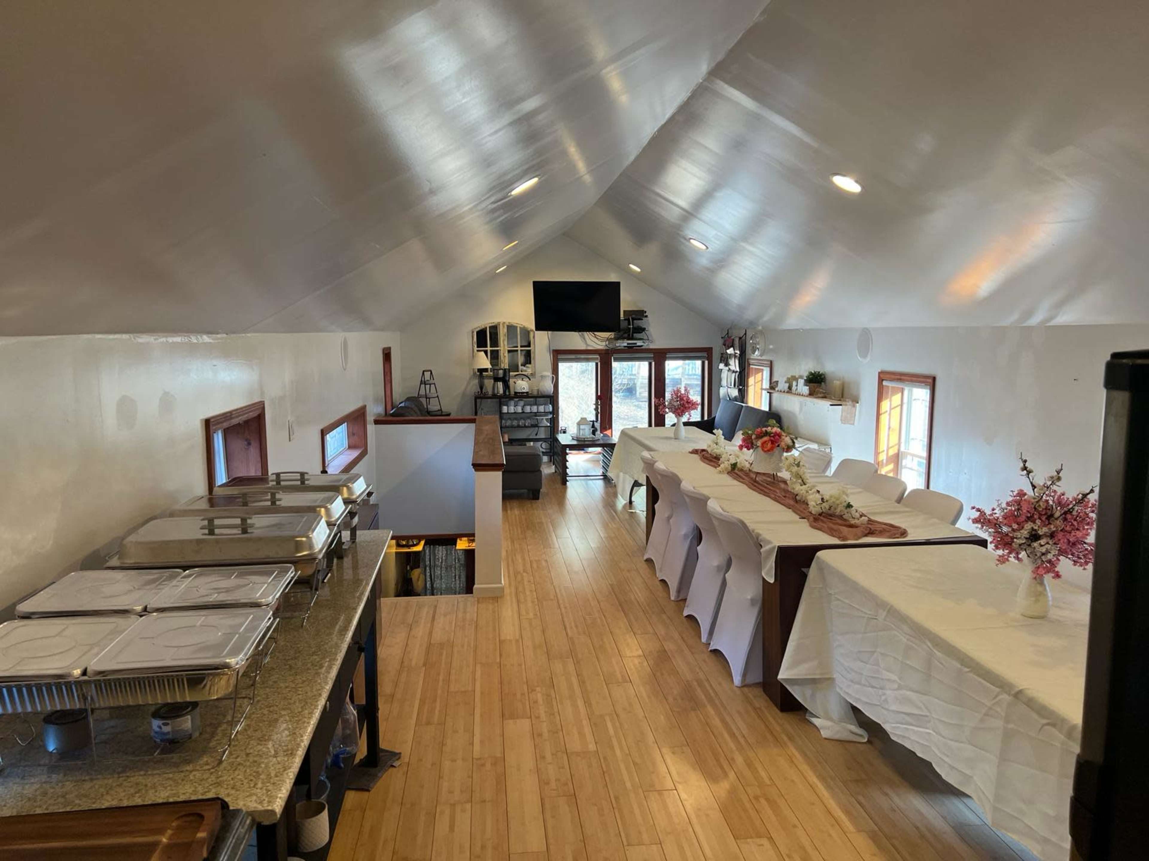 An interior space with tables laid out for a gathering, featuring a buffet setup and a seating area with large windows.