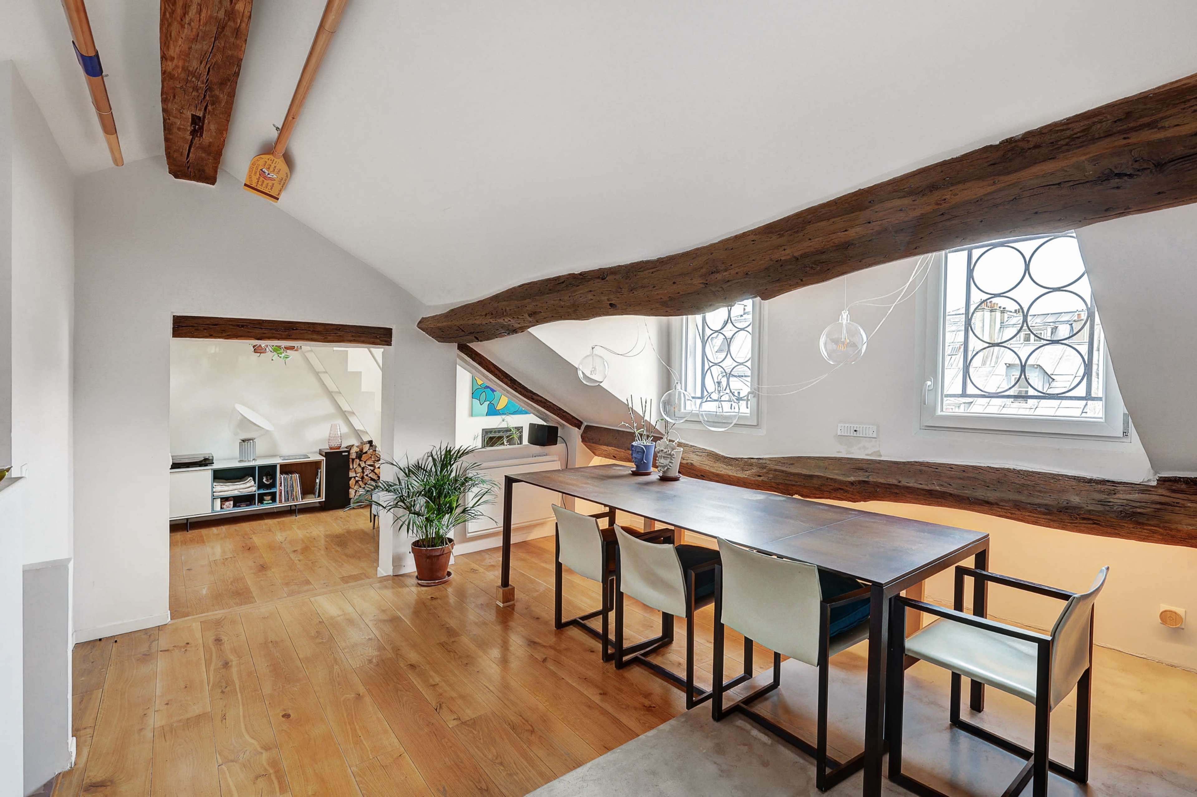 A dining area with a long wooden table and chairs beneath exposed wooden beams and large windows.