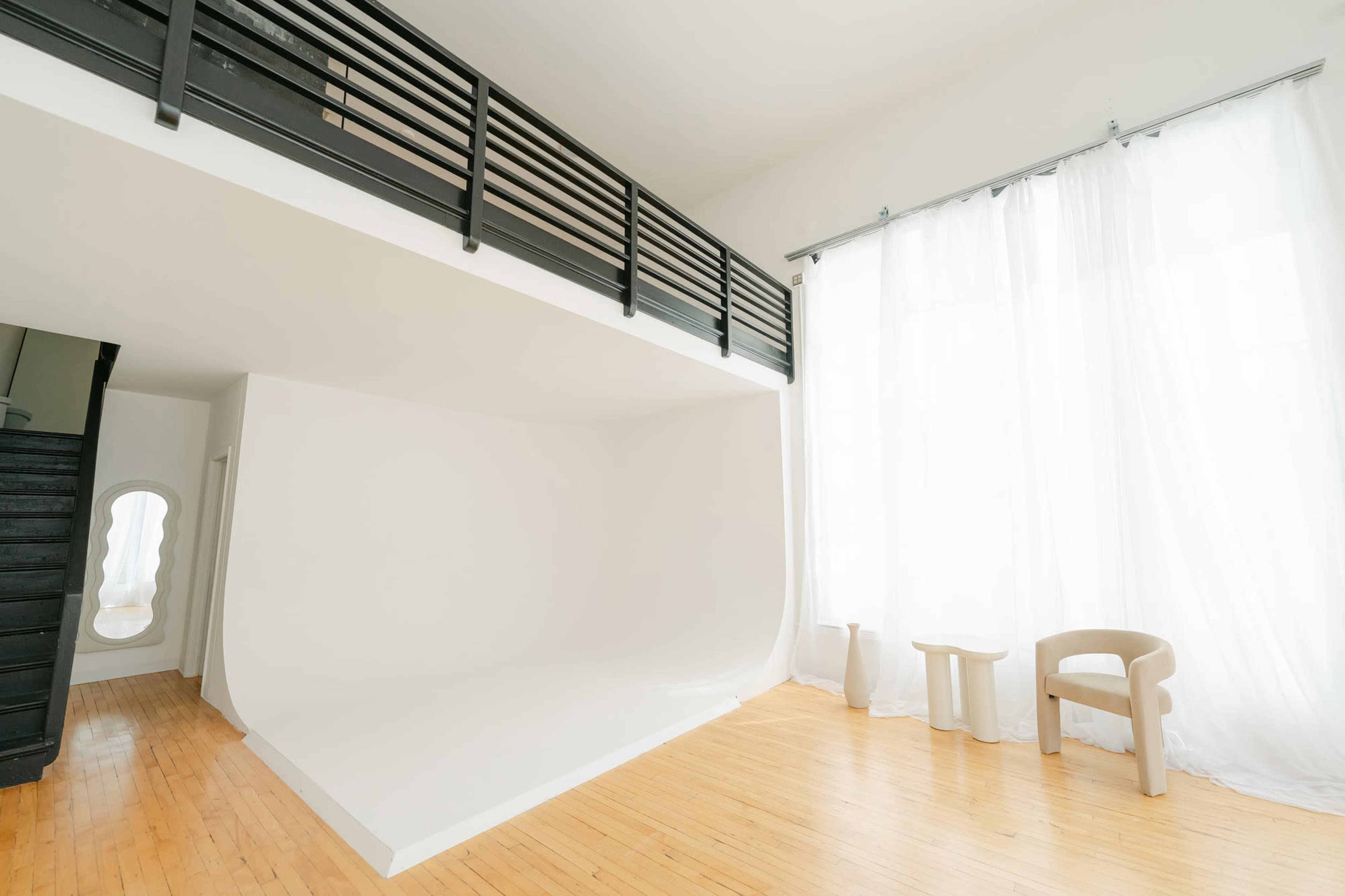 A spacious interior with a large seamless white wall, wooden flooring, and soft natural light filtering through sheer curtains.