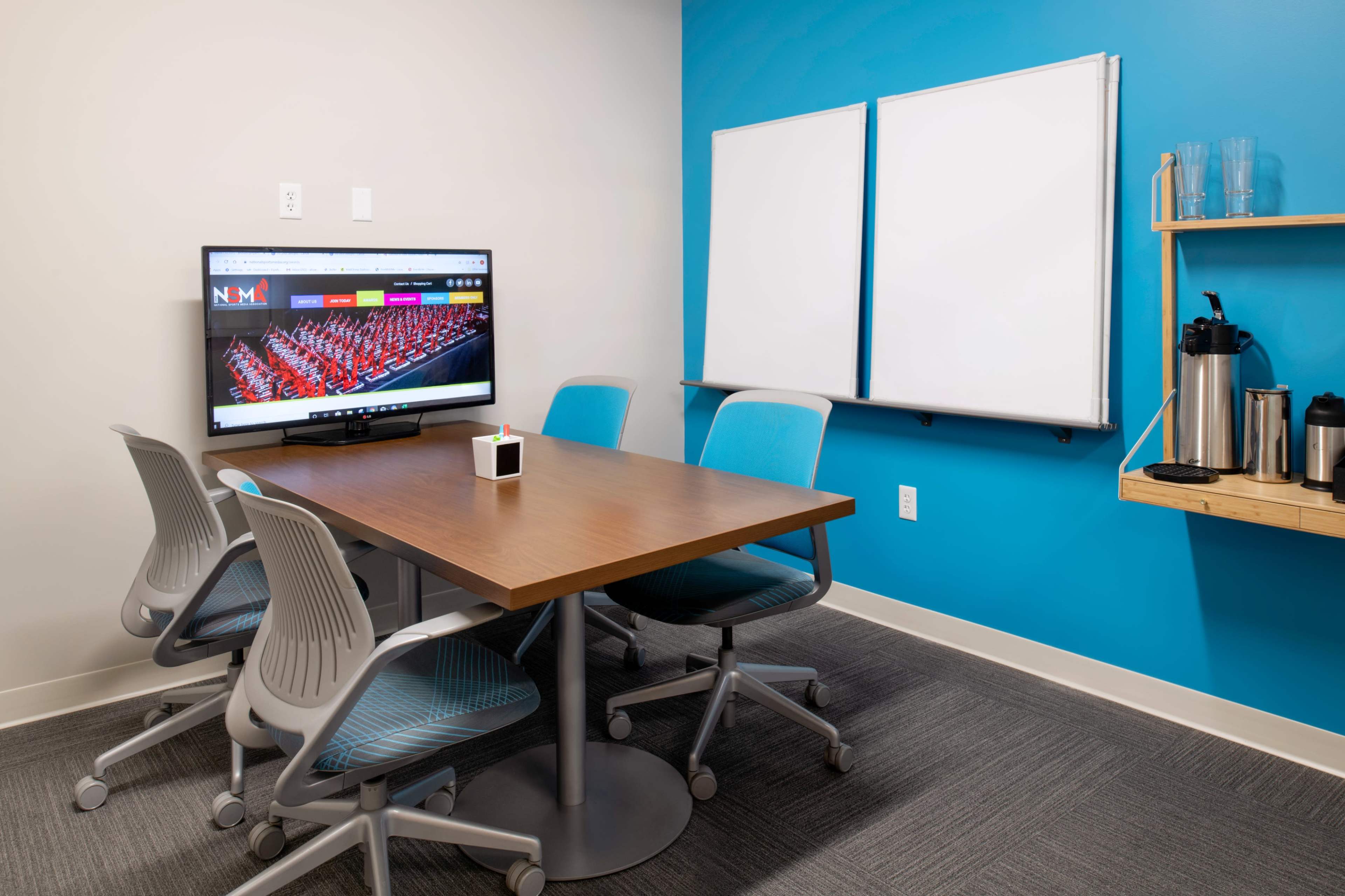 A small meeting room with a wooden table, four chairs, a television displaying a website, and a pair of whiteboards on the wall.