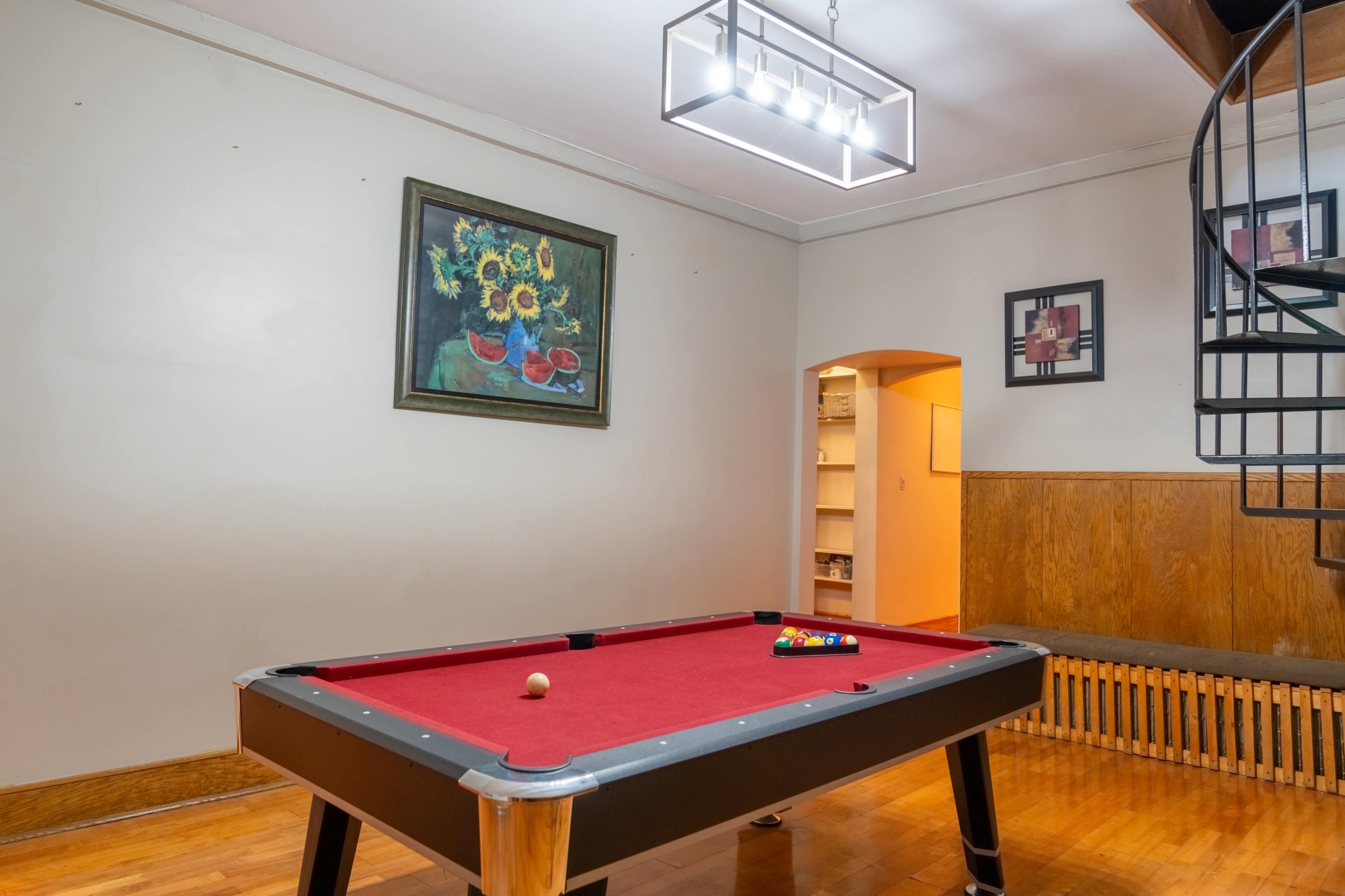 A room with a red pool table in the center, a spiral staircase on the right, and a painting of sunflowers on the wall.