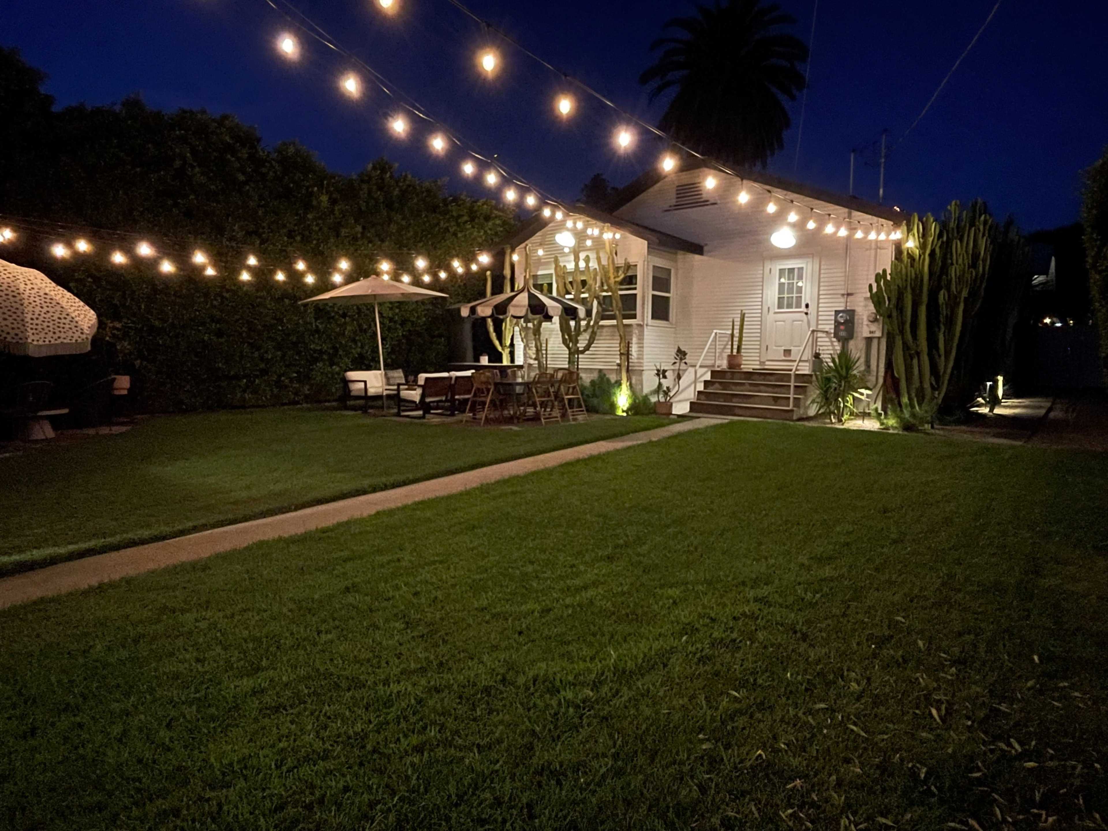 A well-lit backyard features string lights above a green lawn, leading to a cozy house surrounded by cacti and outdoor furniture.