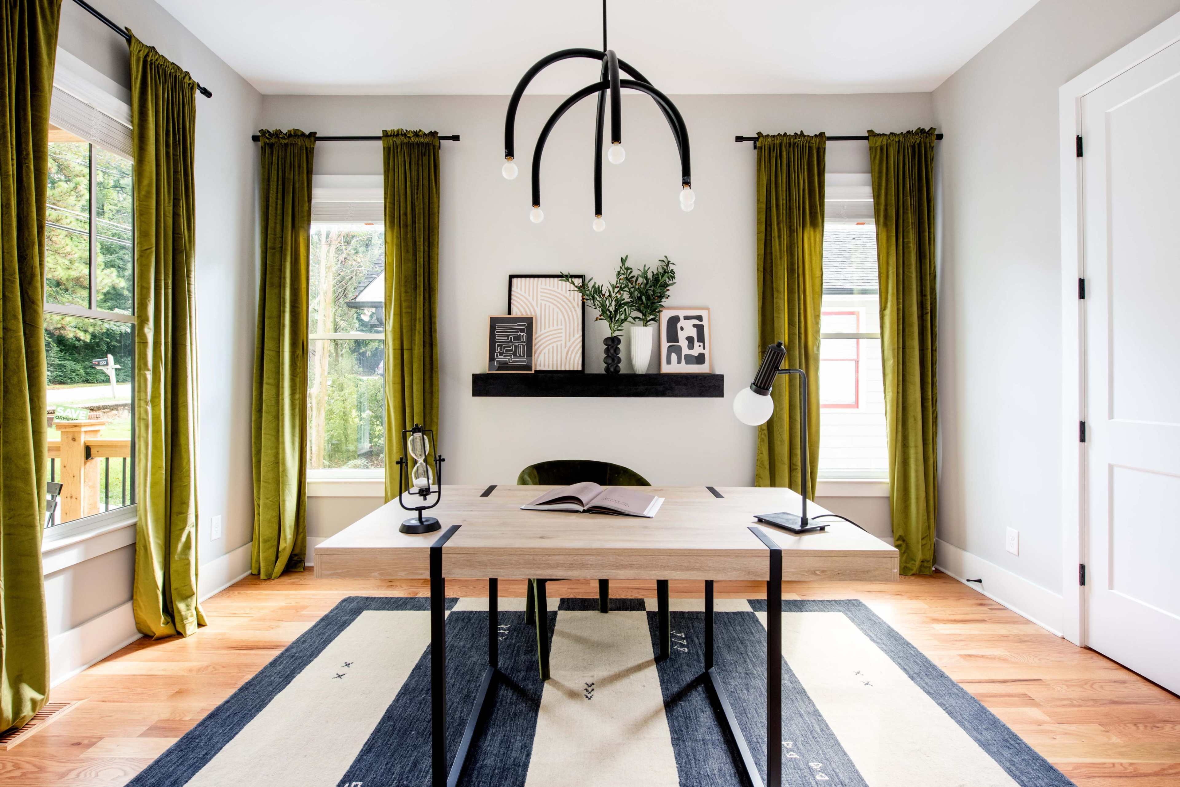 A modern home office with a wooden desk, two desk lamps, a wall shelf with decorative items, and large windows dressed in green curtains.