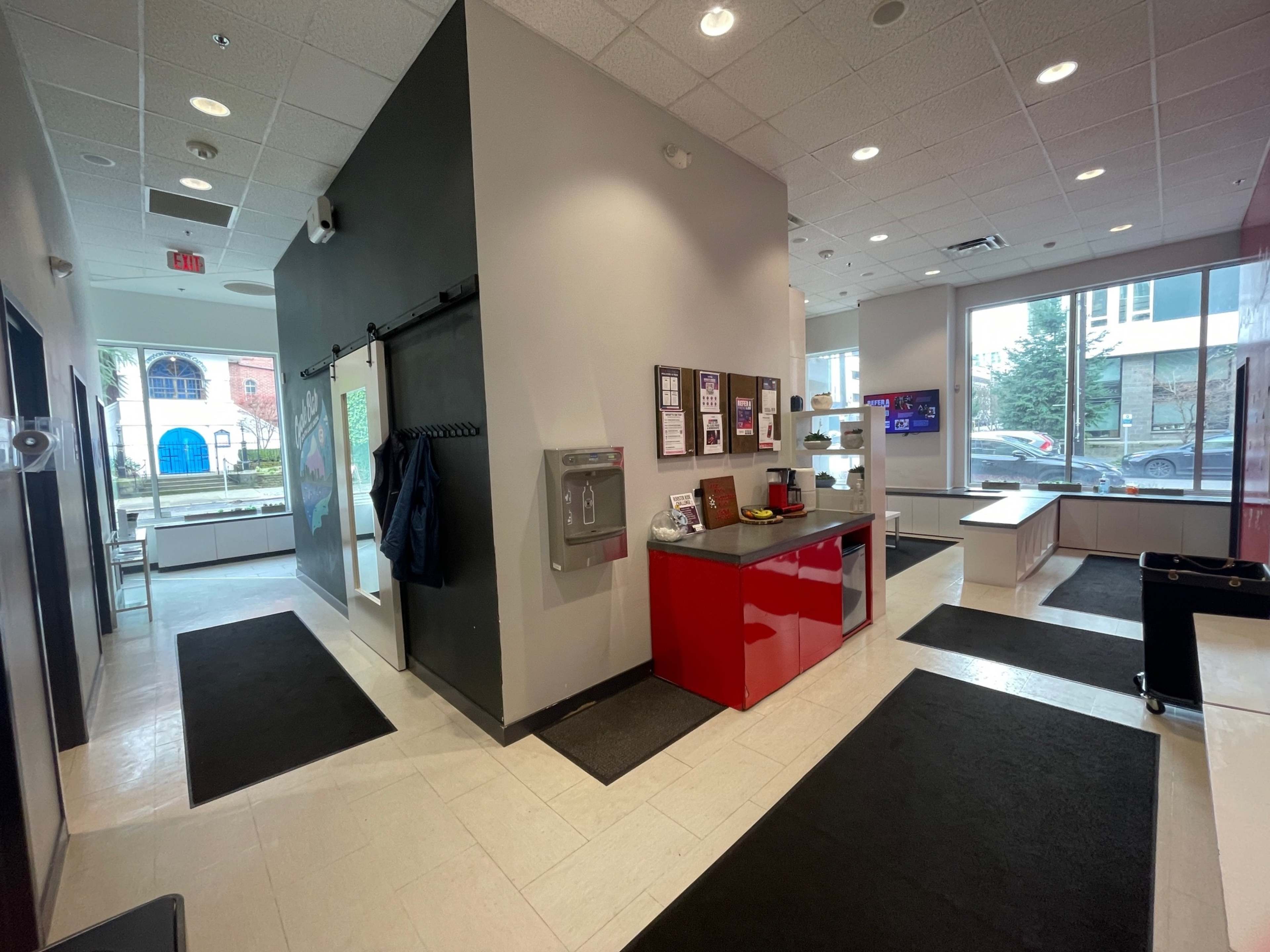 The image shows a modern lobby area with a reception counter, black mats on the floor, and large windows letting in natural light.