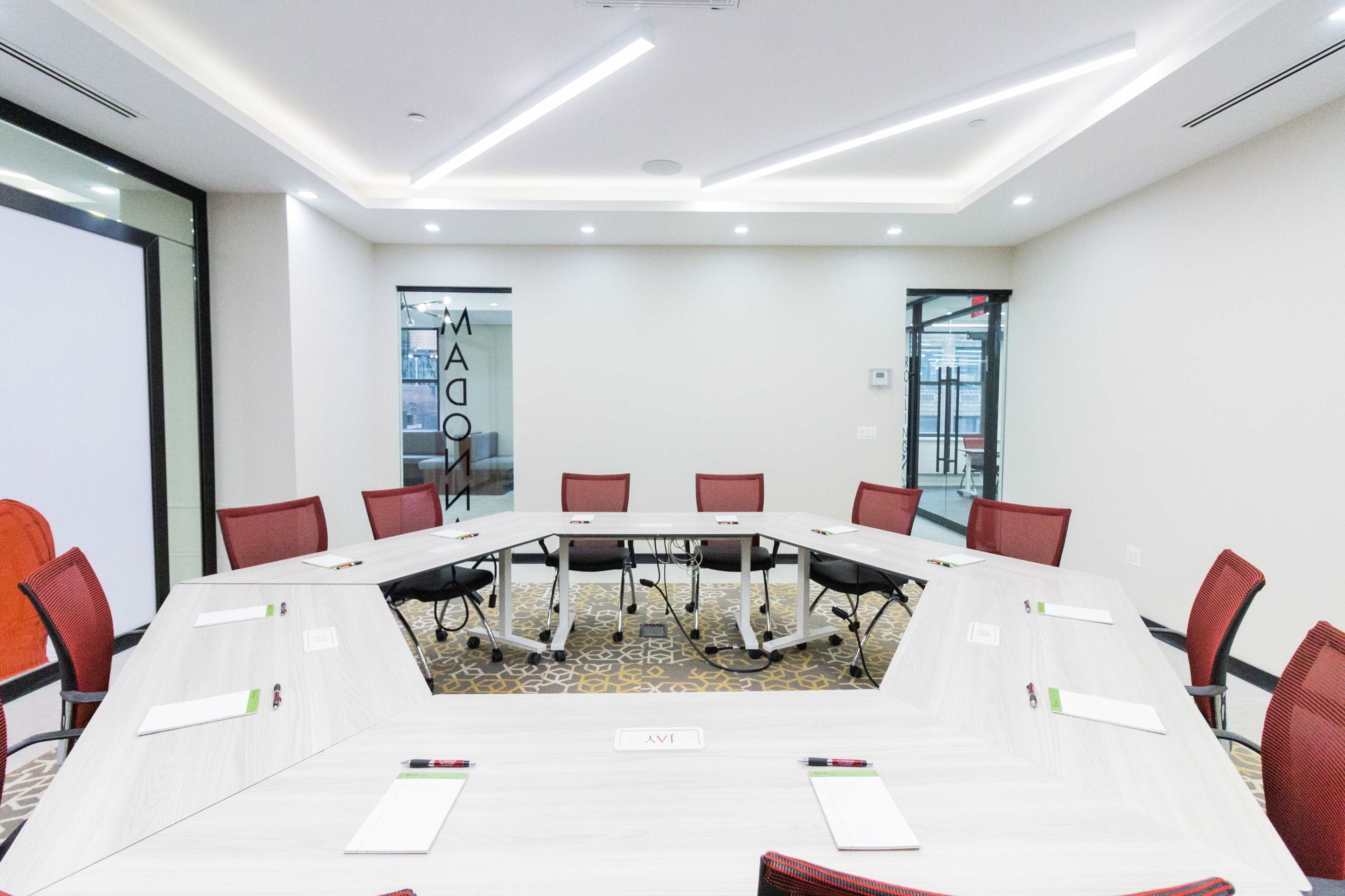 A modern conference room features a hexagonal table surrounded by red chairs, with a glass wall displaying the word "MAGON."