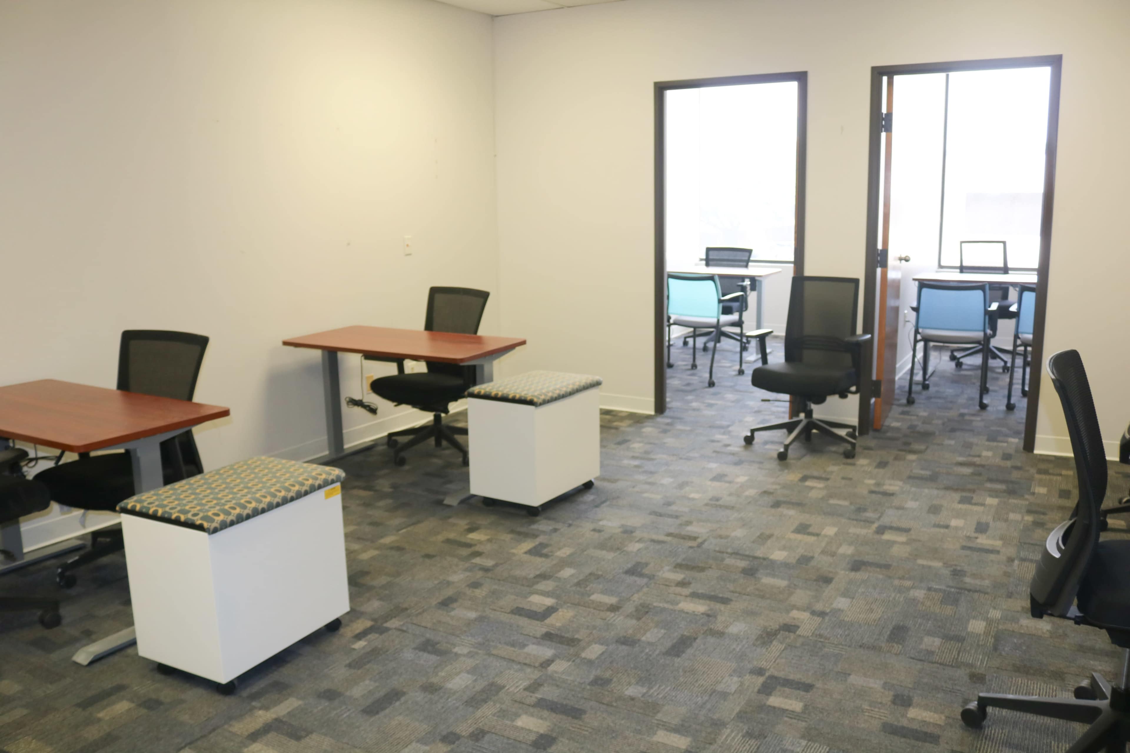 The image shows a modern office space with two desks and seating in an open layout, featuring a neutral color scheme and light carpeting.