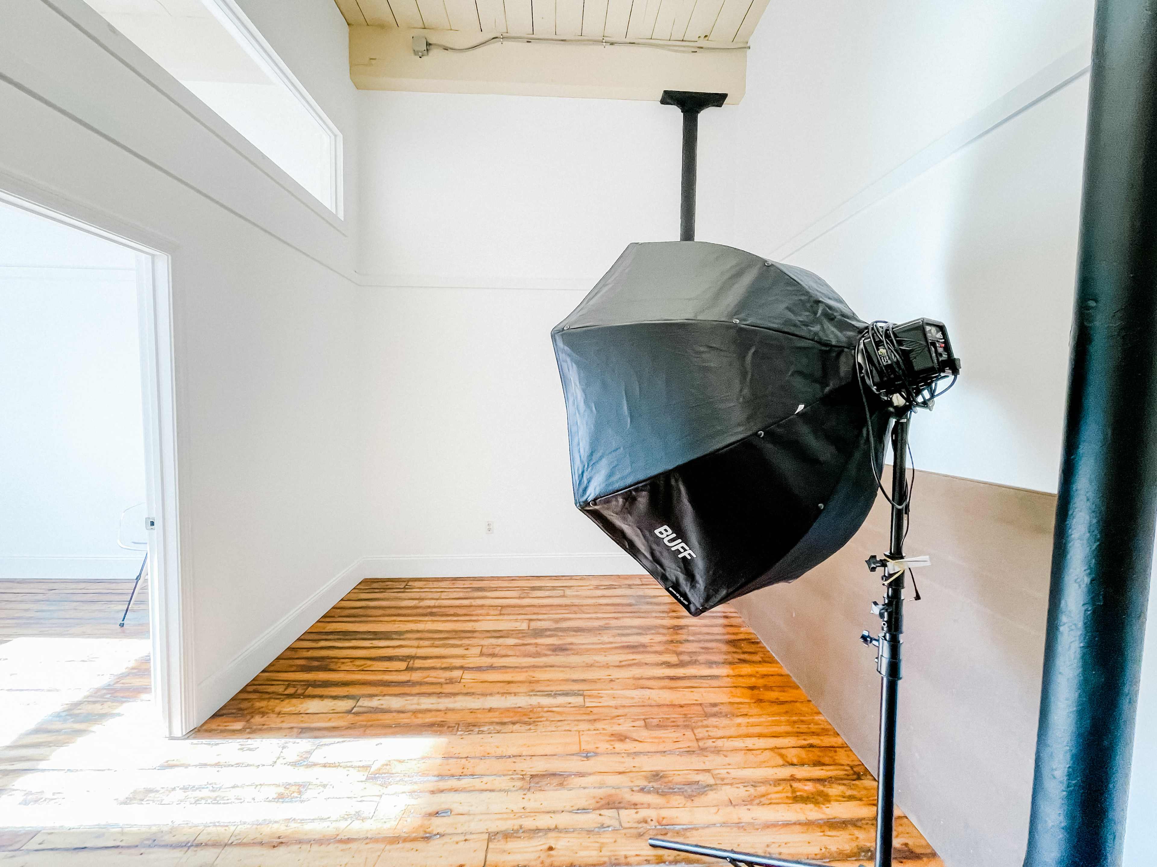 A large softbox light stands on a stand in an empty room with wooden floors and white walls.