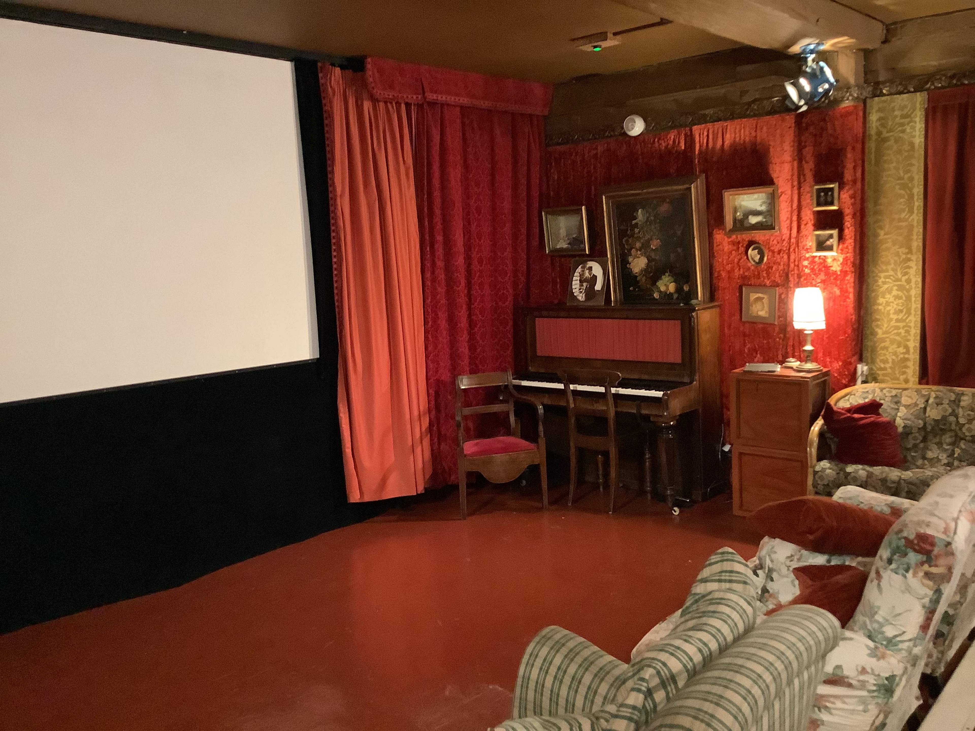The room features a large projector screen, a piano with a chair in front, a small lamp on a cabinet, and a patterned sofa against the wall.