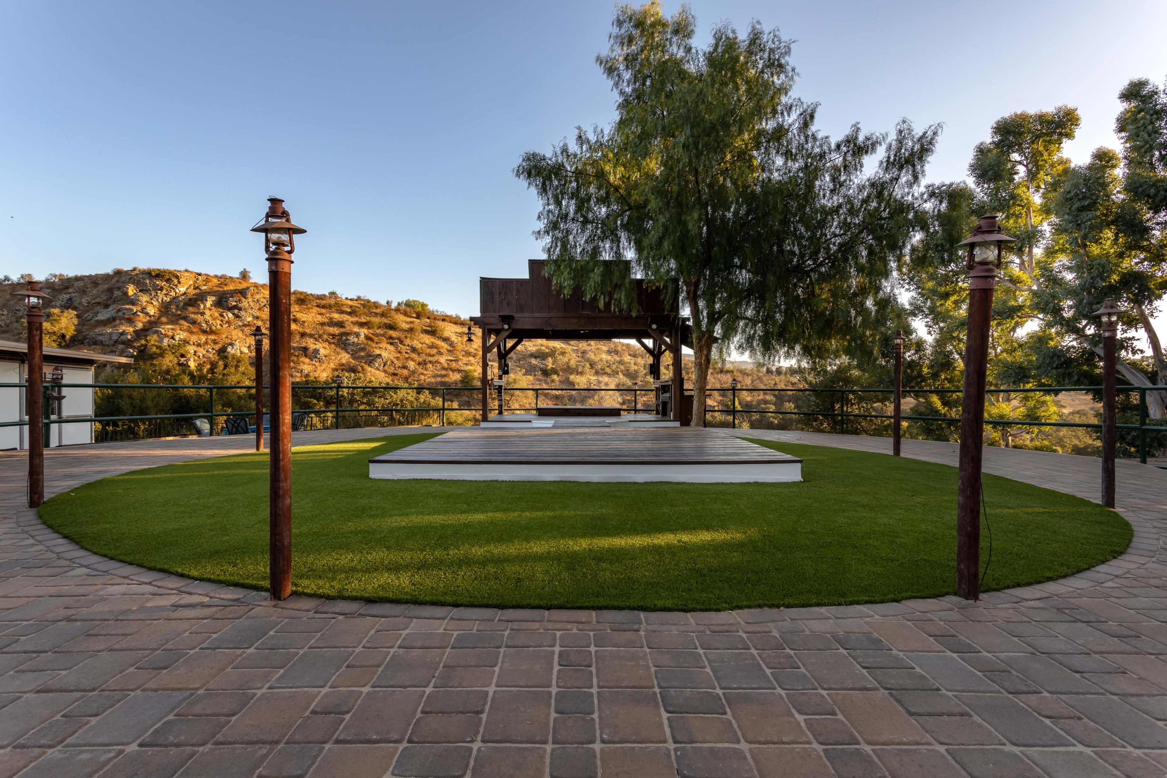 The scene shows a circular grassy area with a wooden platform and a gazebo, surrounded by stone paving and trees on a hillside.