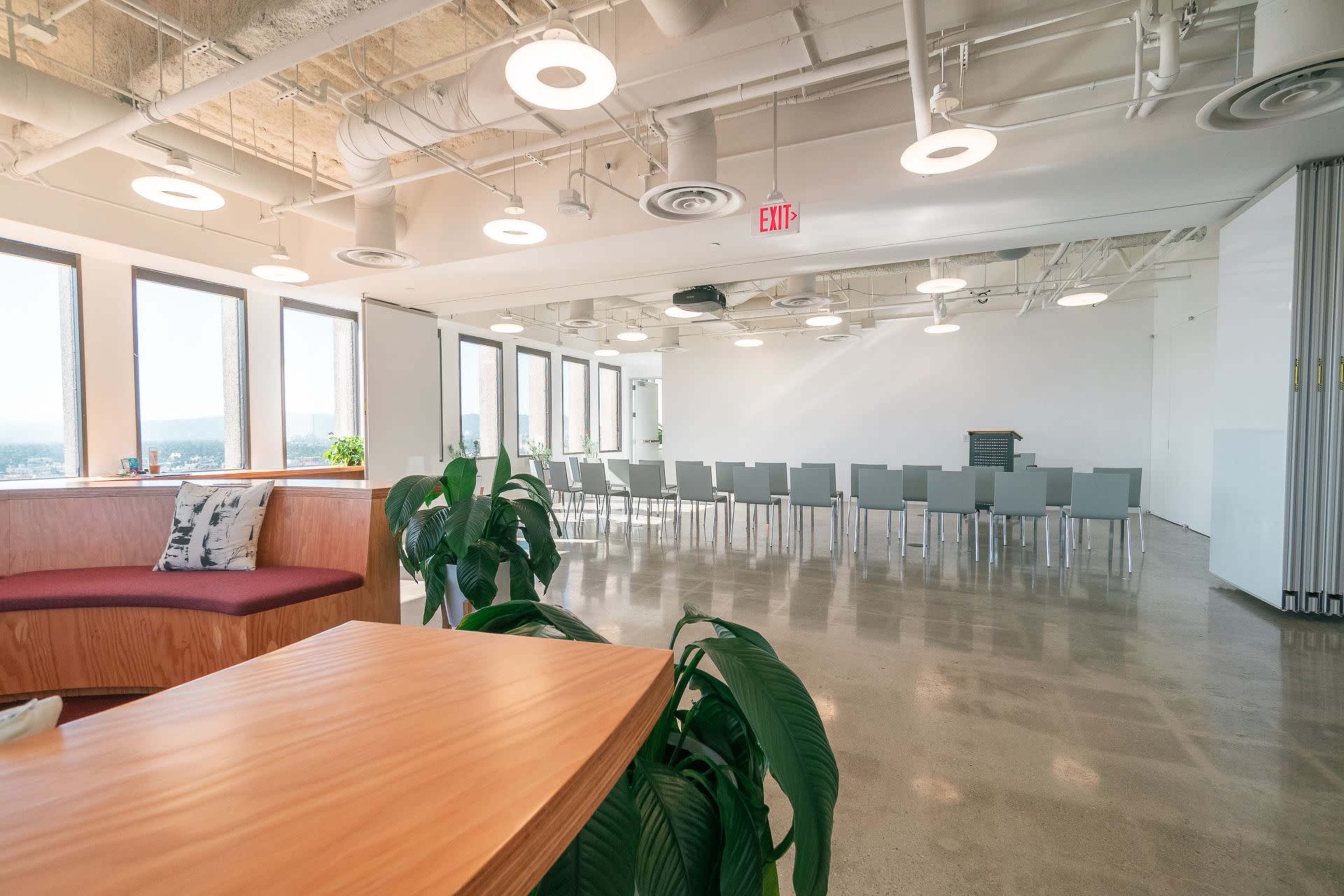 A modern office space with large windows, a meeting area featuring a long table and chairs, and a seating area with a wooden table and plants.