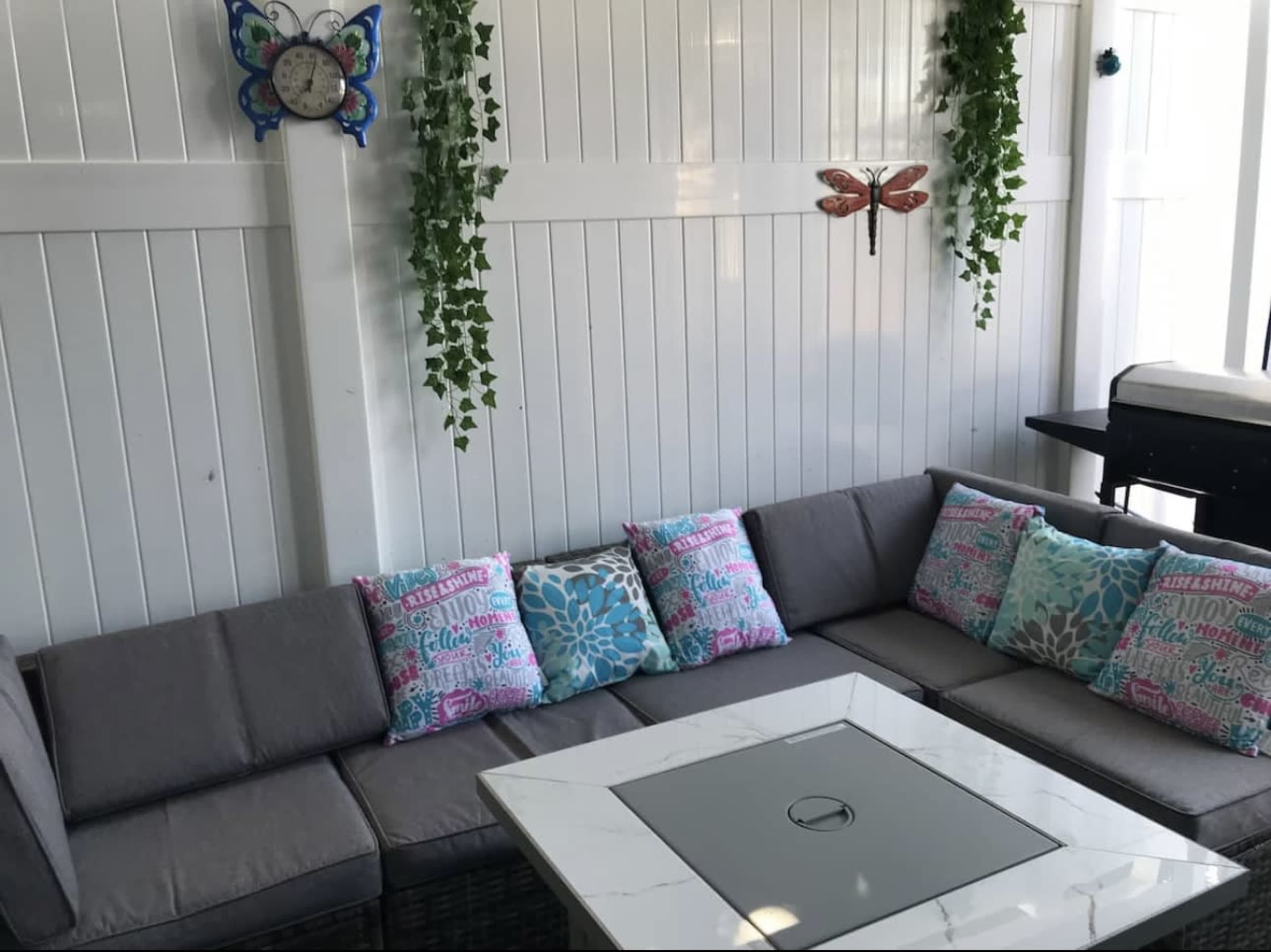 The image shows a cozy seating area with a sectional sofa adorned with colorful throw pillows, a square coffee table, and decorative wall elements including a clock and wall hangings.