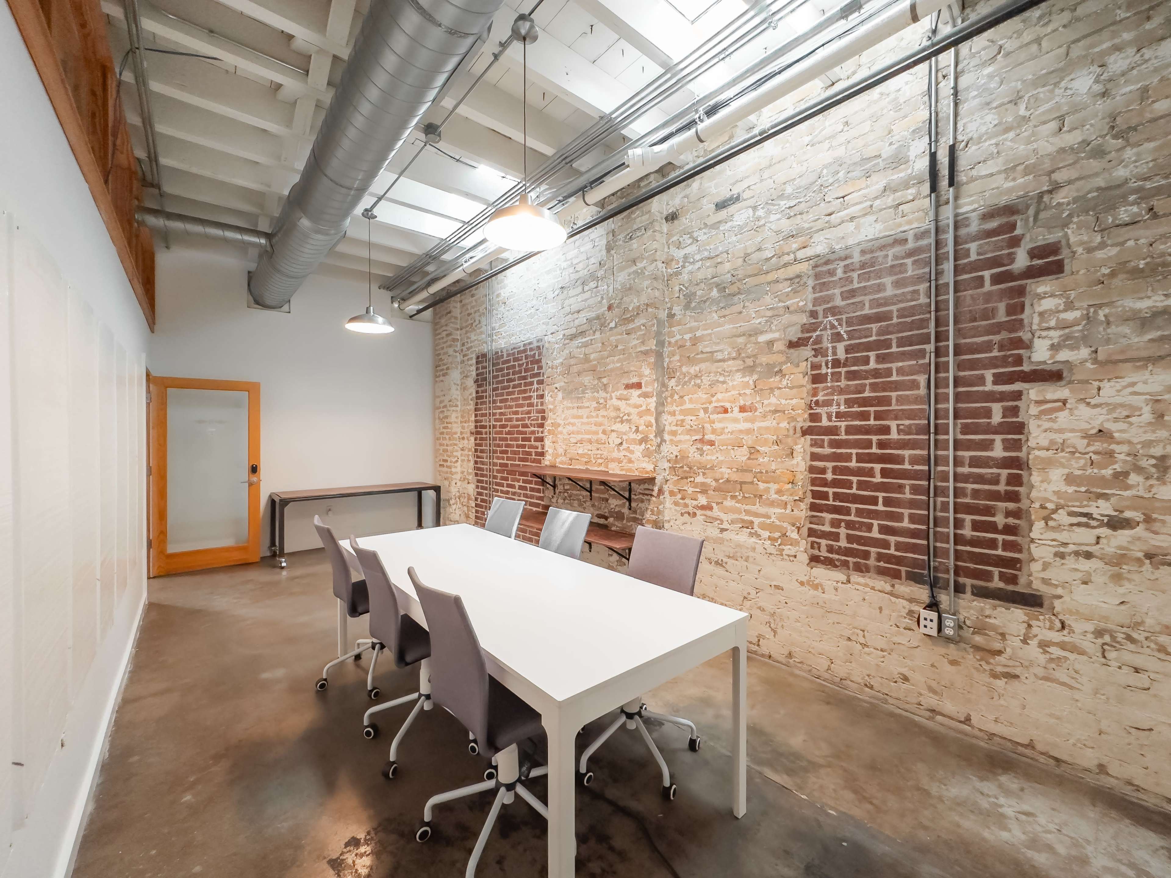 A minimalist conference room with a white table and six chairs, featuring exposed brick walls and industrial lighting.