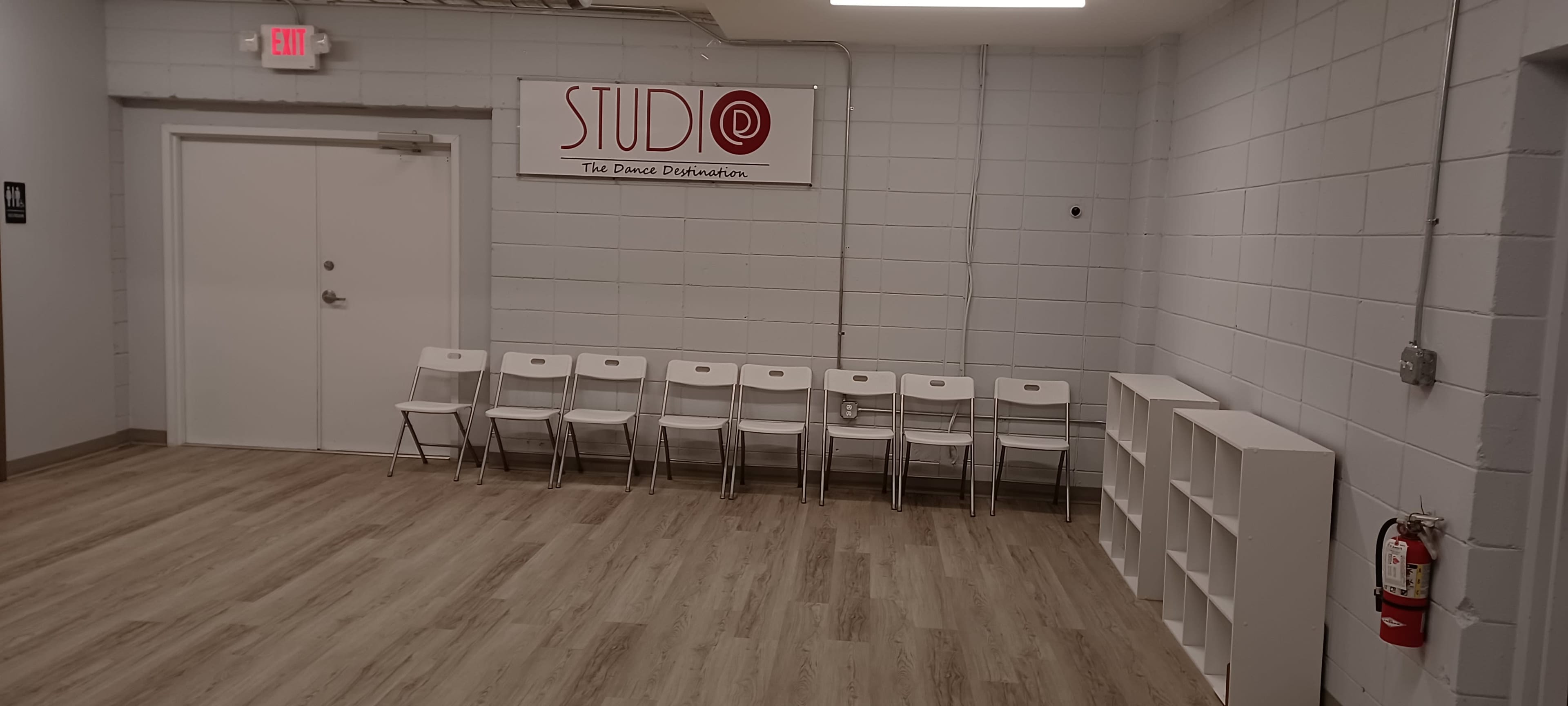 A series of white chairs are lined up against a gray wall in a spacious room featuring a sign that reads "STUDIO - The Dance Destination."
