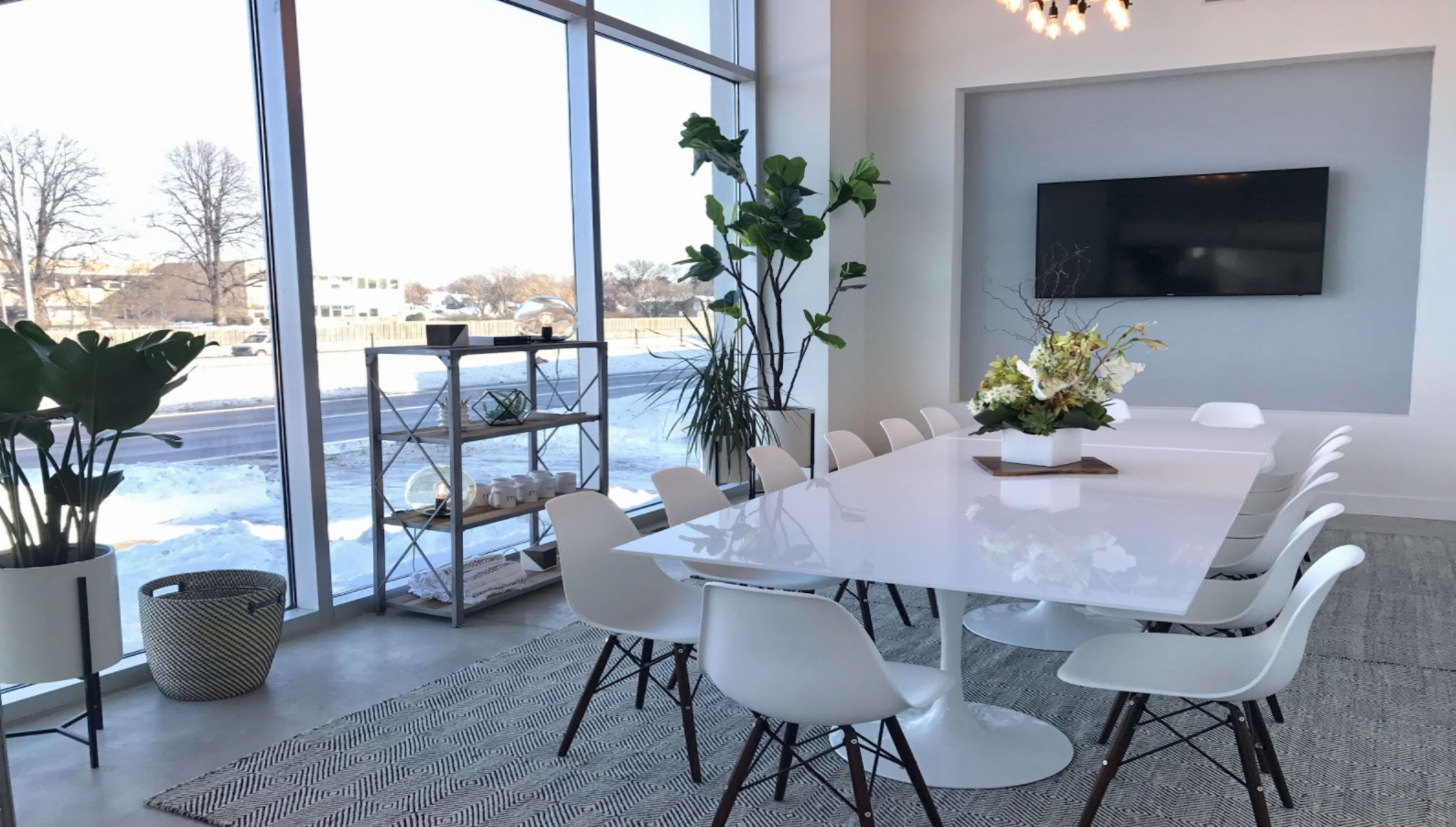 A modern conference room features a long white table surrounded by white chairs, large windows with a view of outdoors, and a decorative plant arrangement.