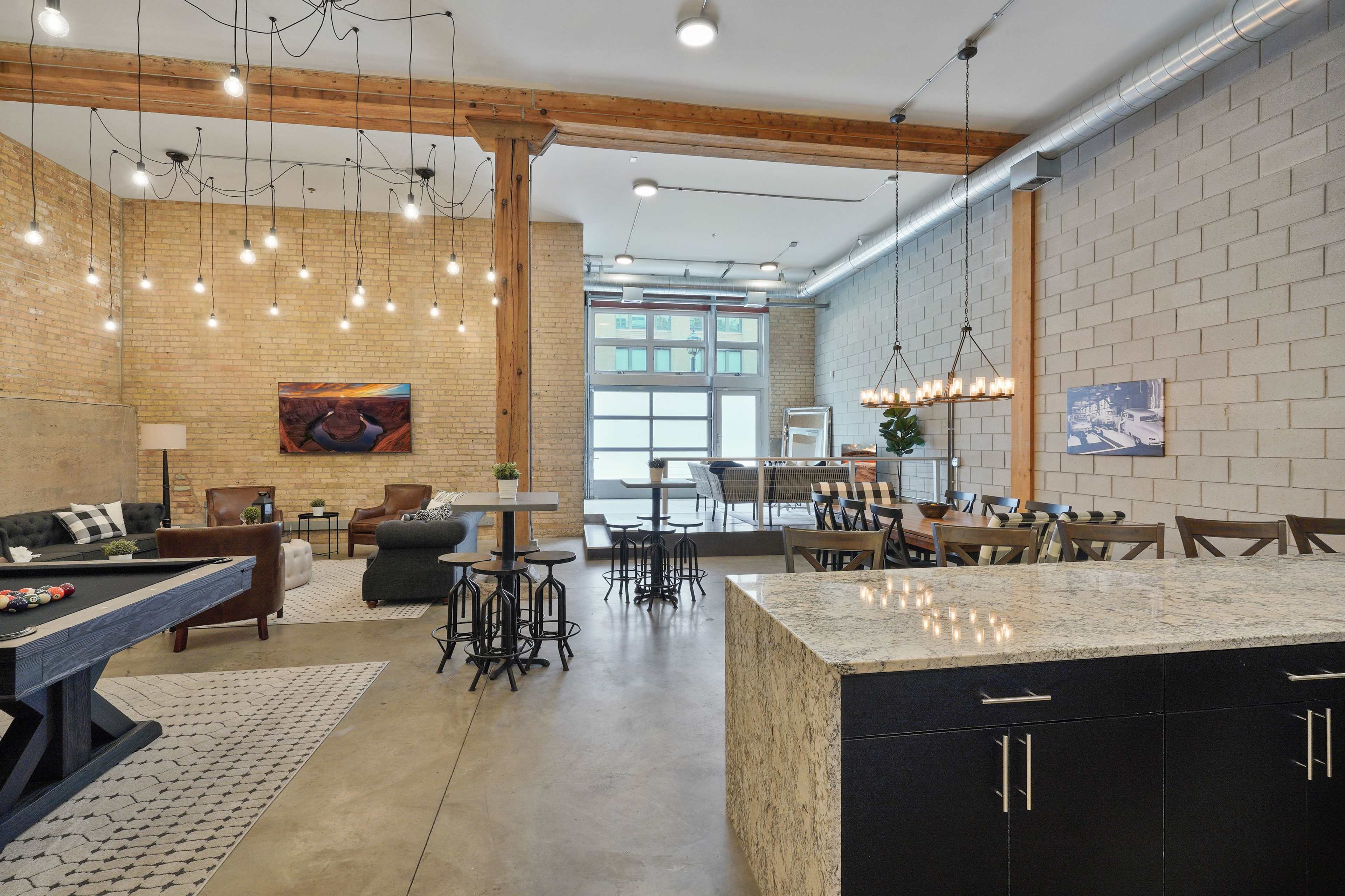 A spacious industrial-style interior with a kitchen island, dining area, lounge seating, and a pool table, illuminated by various hanging light fixtures.