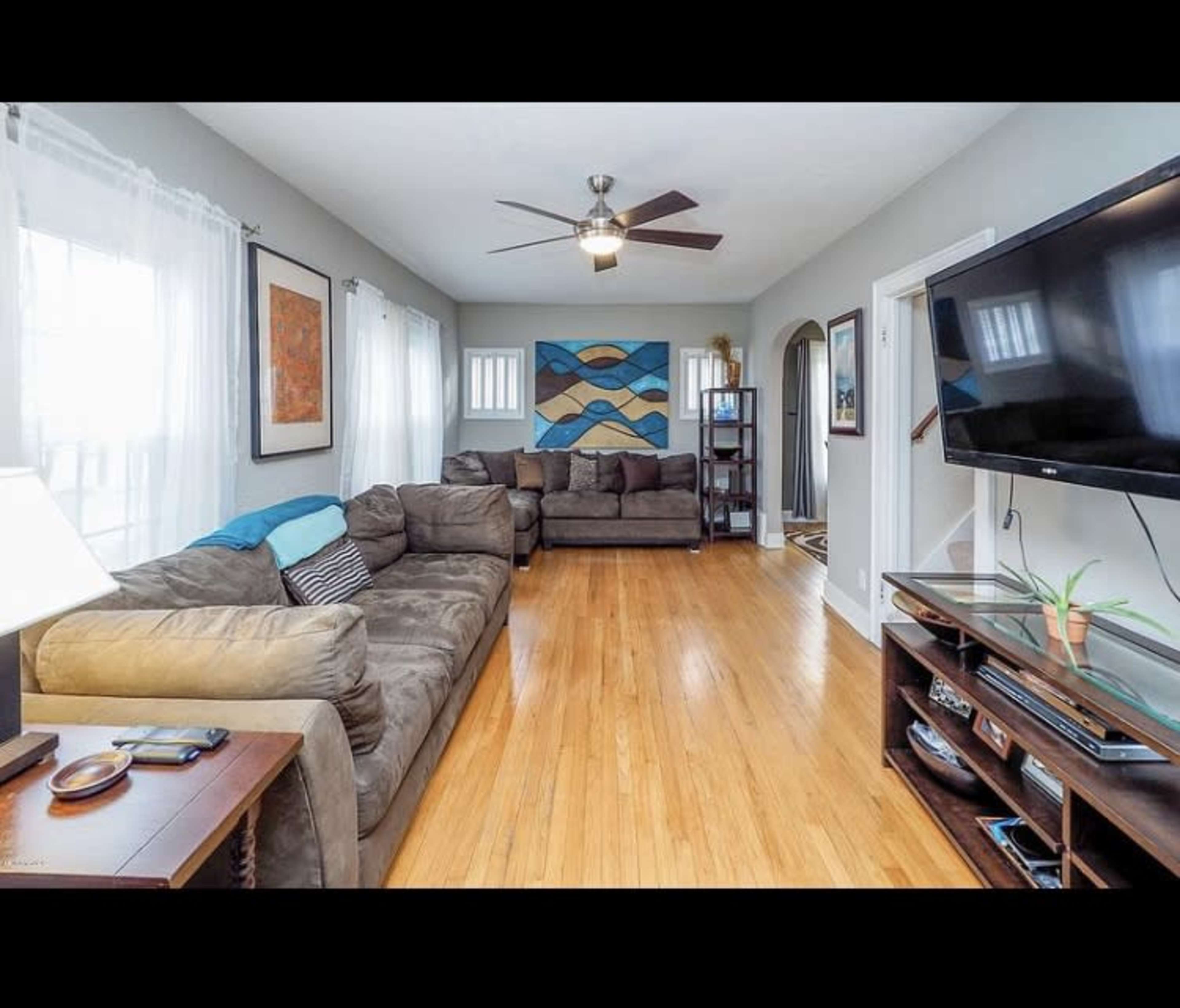 The living room features two large sofas, a coffee table, and a flat-screen TV against a wooden floor, with large windows providing natural light.