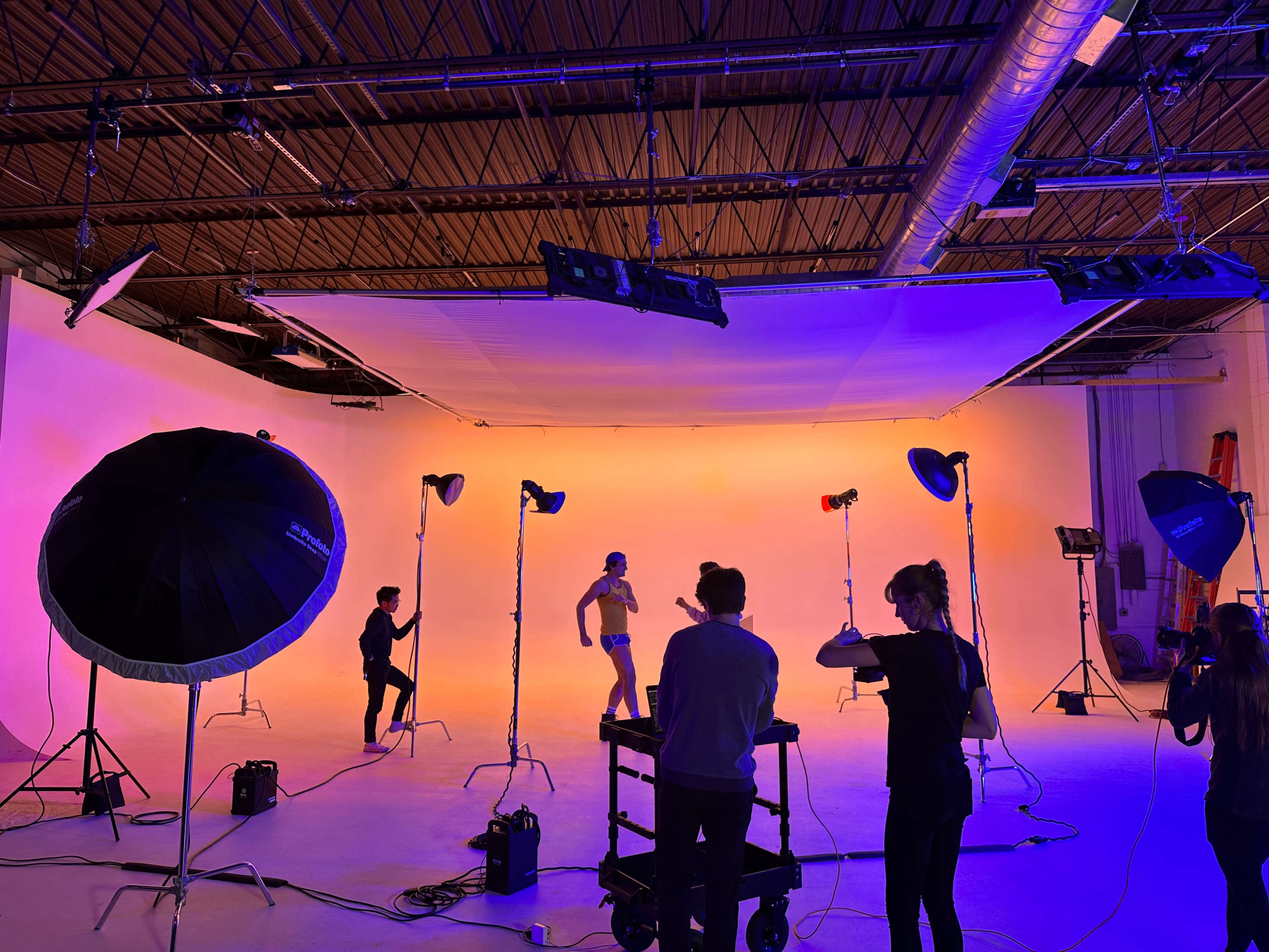 A studio space is set up for a photoshoot, featuring lighting equipment, a backdrop, and several individuals posing and adjusting settings.