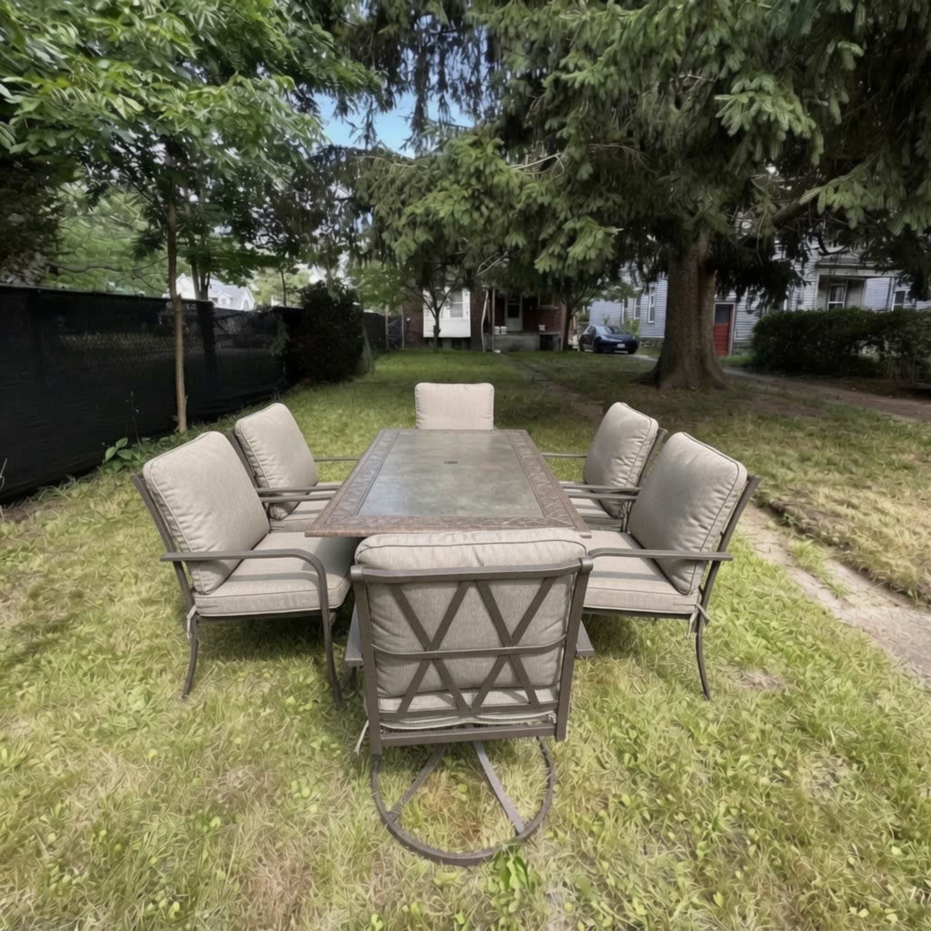 A rectangular dining table surrounded by six cushioned chairs is situated on a grassy area under tall trees.