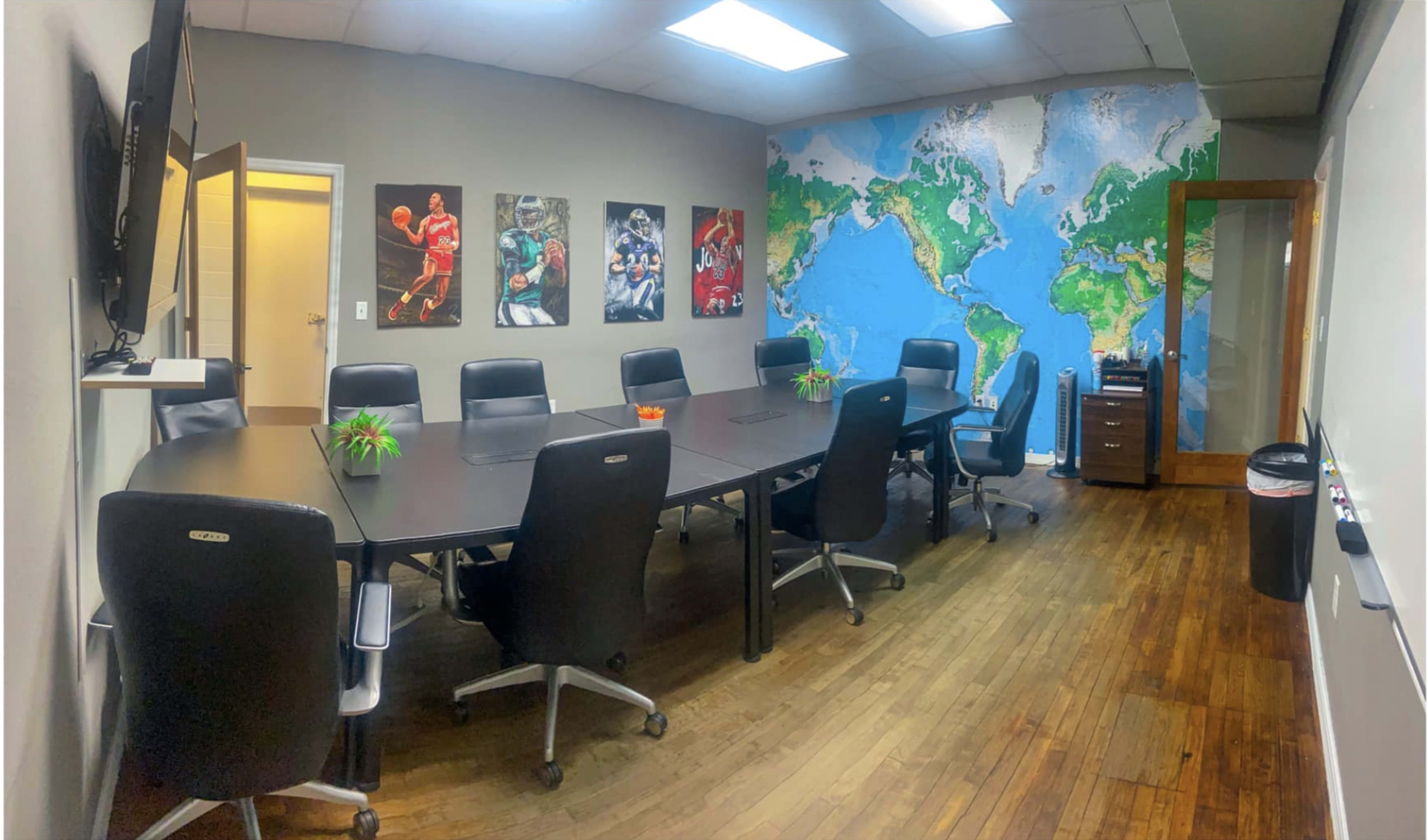 A conference room is furnished with a large table surrounded by black chairs, featuring a world map mural on one wall and sports-themed artwork on another.