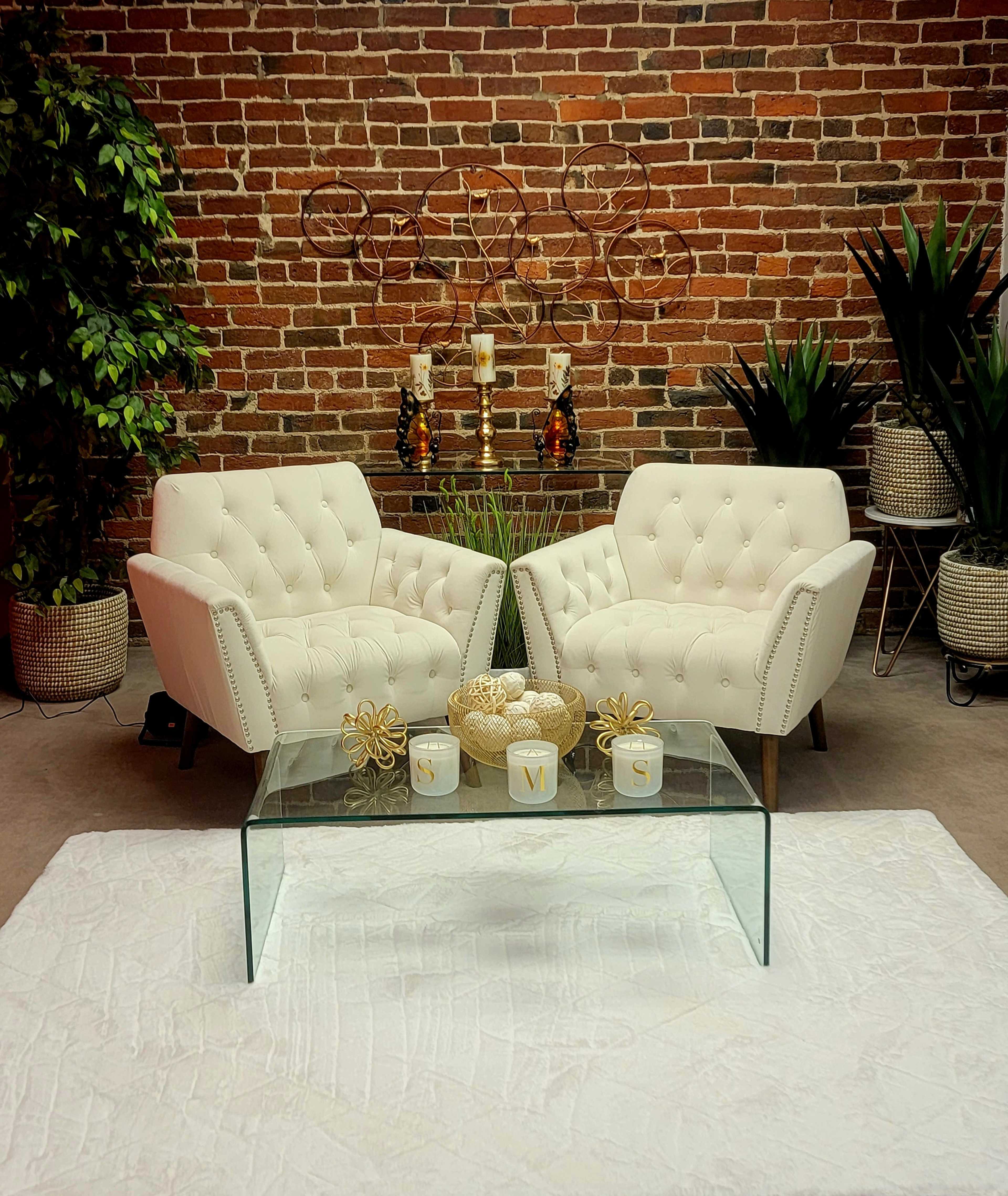 The scene features two white tufted chairs positioned facing each other over a glass coffee table, set against a brick wall decorated with plants and candle holders.