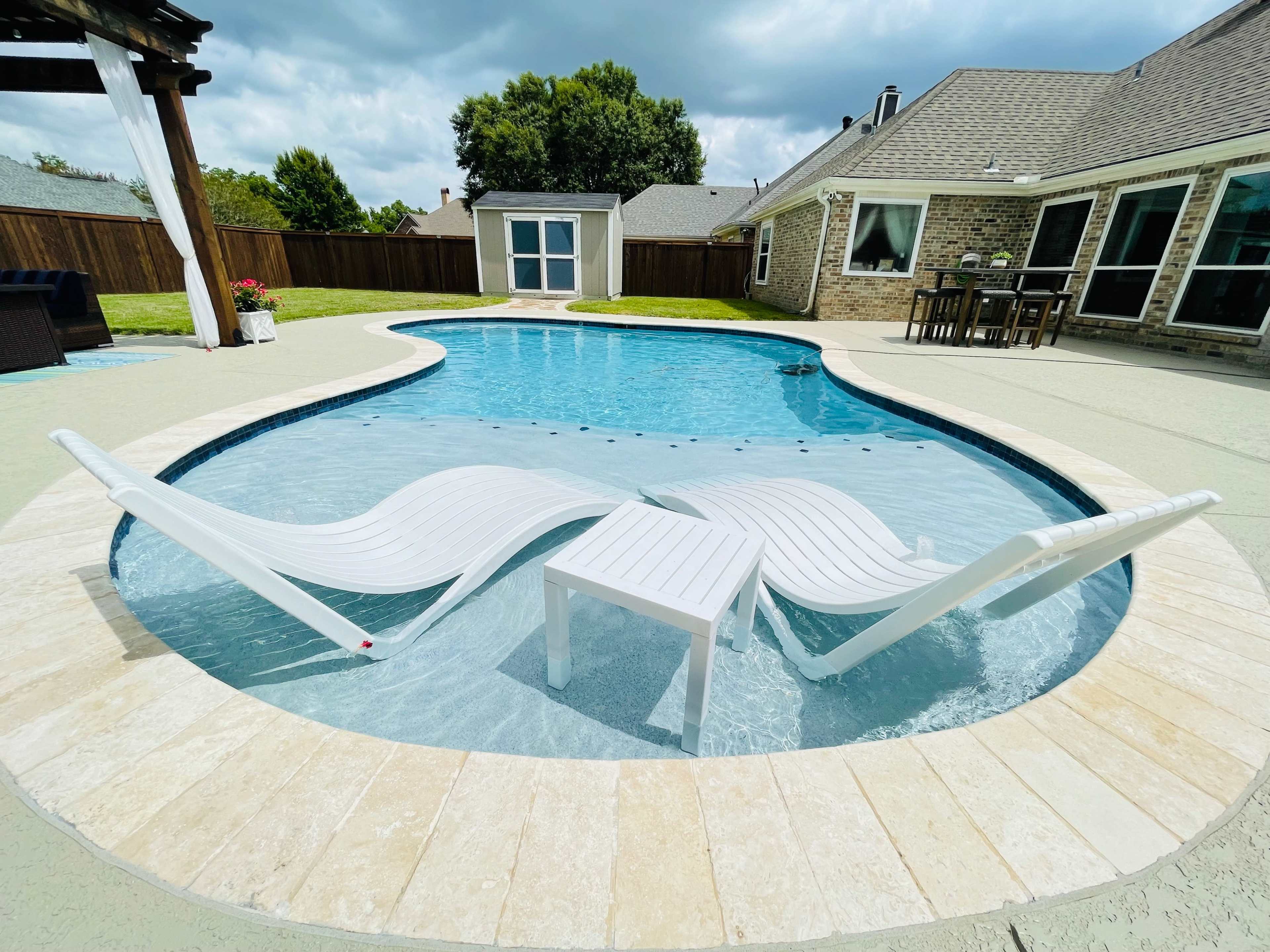 A swimming pool with two white lounge chairs and a small table placed in the shallow water.