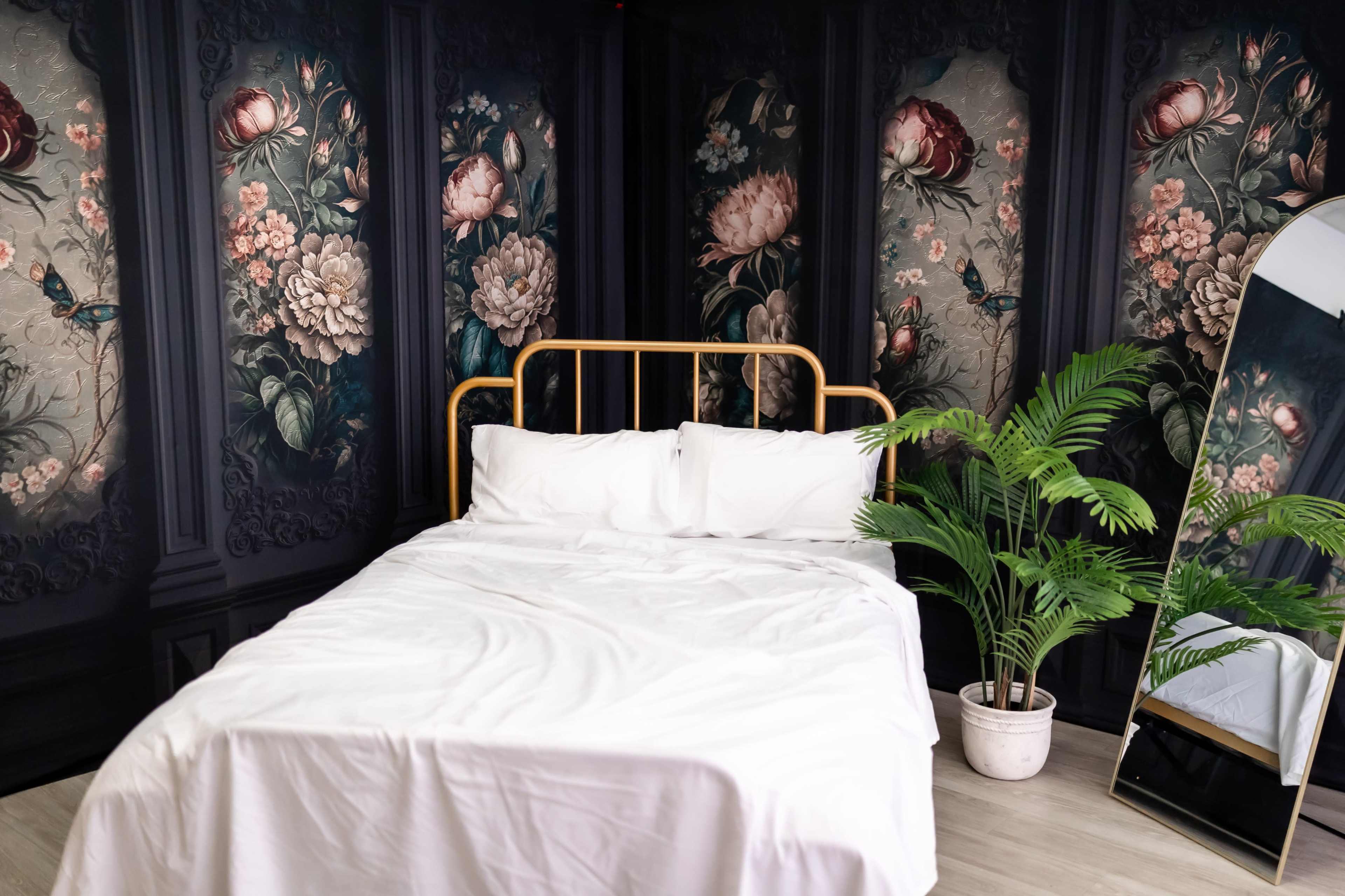 The image shows a bedroom with a gold metal bed frame, white bedding, floral wallpaper, and a large mirror beside a potted plant.