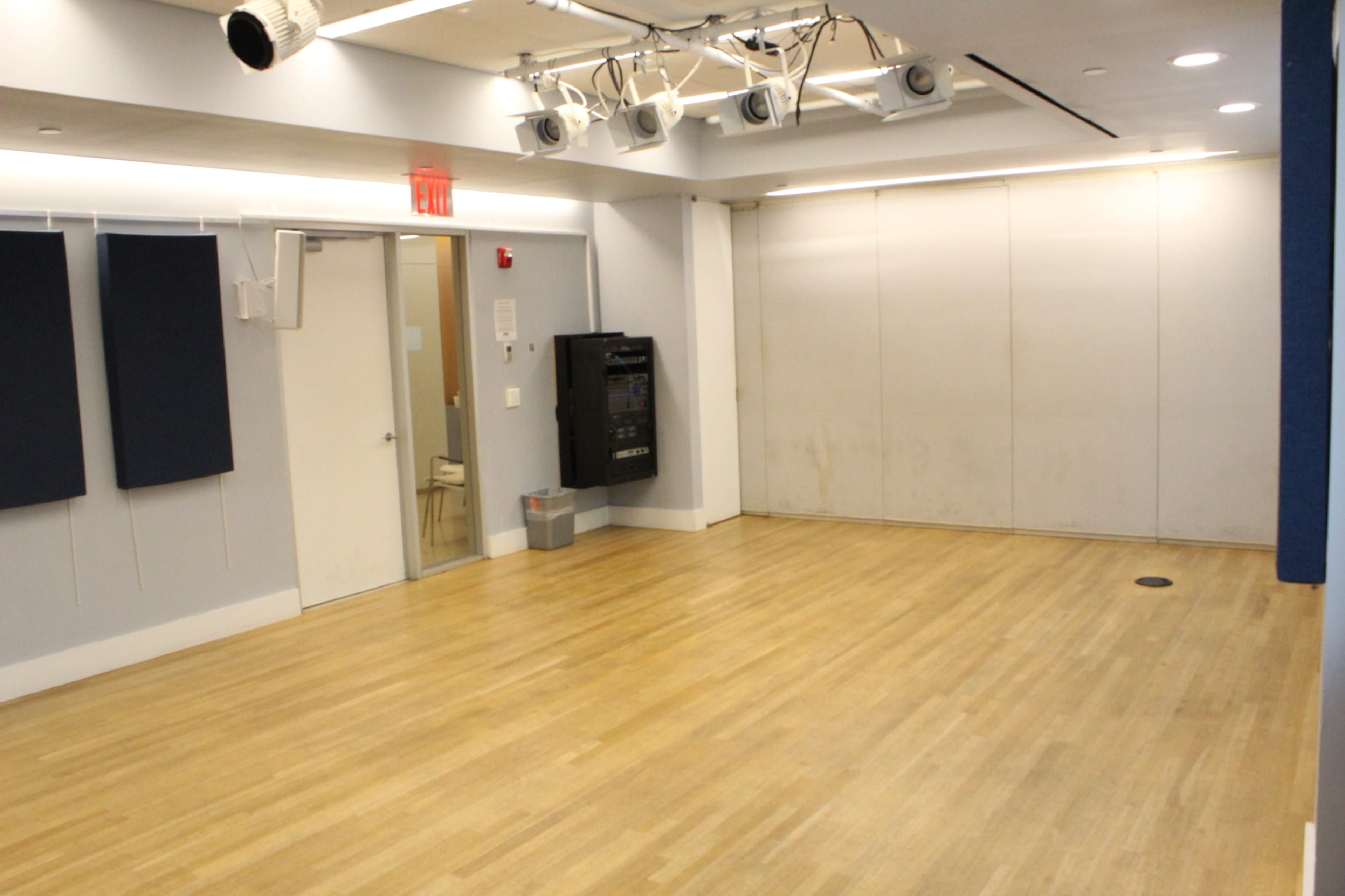 The image shows a vacant room with wooden flooring, a soundproofing panel on the left wall, and several ceiling-mounted spotlight fixtures.