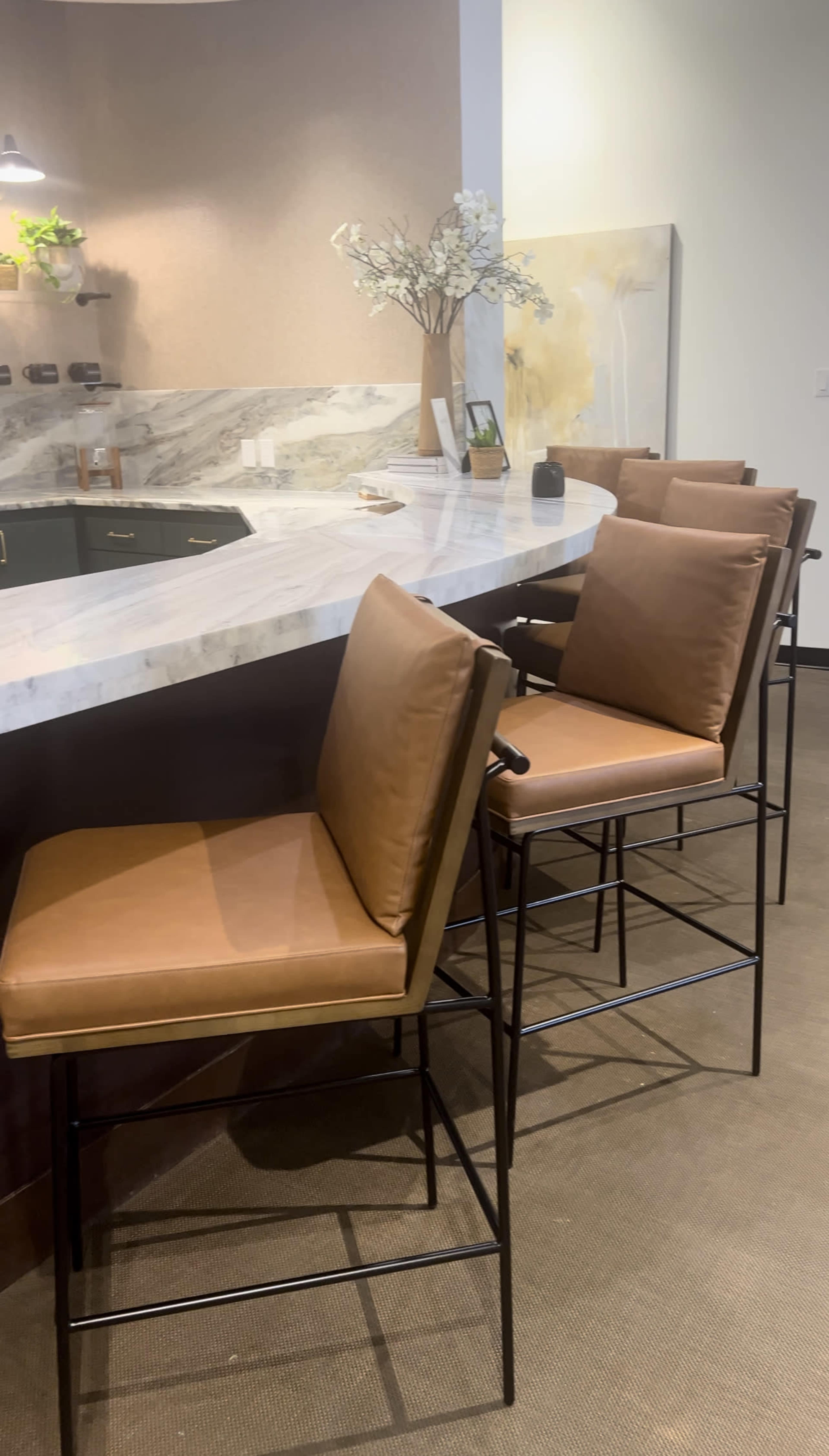 A modern kitchen bar area features sleek metal stools with tan cushions arranged around a marble countertop.