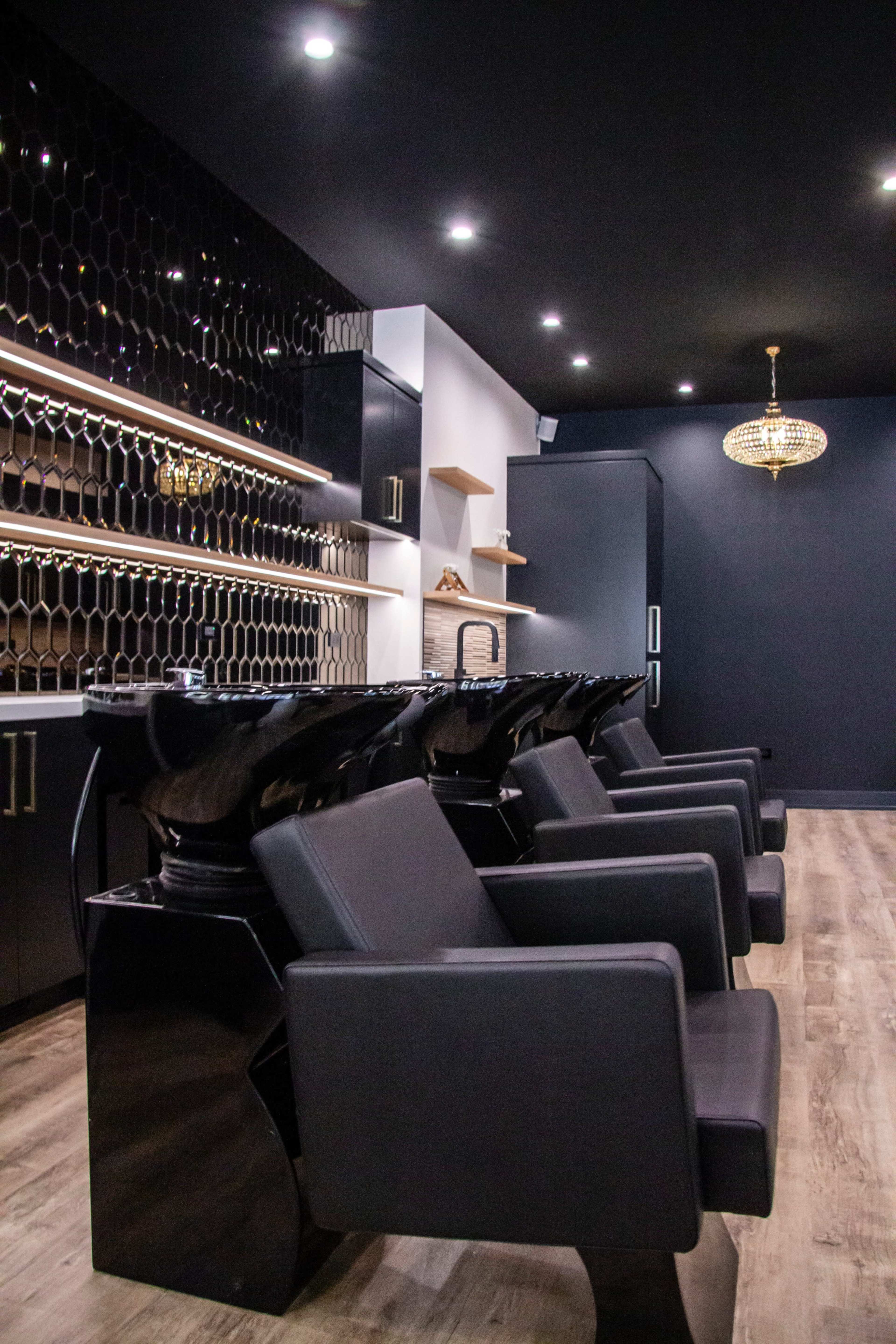 The image shows a modern hair salon interior with black shampoo bowls and sleek black chairs arranged in a row against a dark wall with stylish lighting.