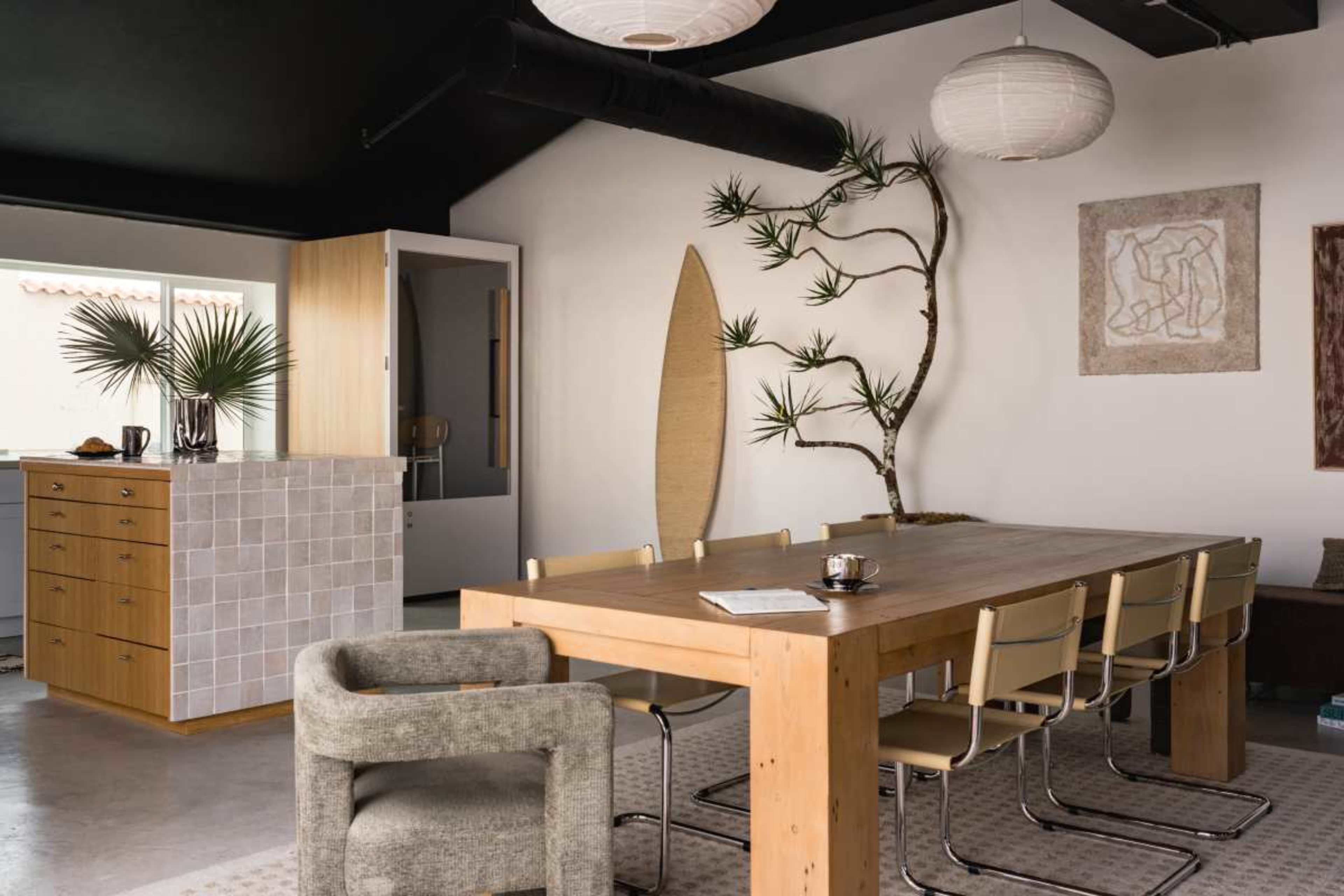 The image features a modern dining area with a large wooden table surrounded by minimalist chairs, complemented by a geometric kitchen island and decorative plants.