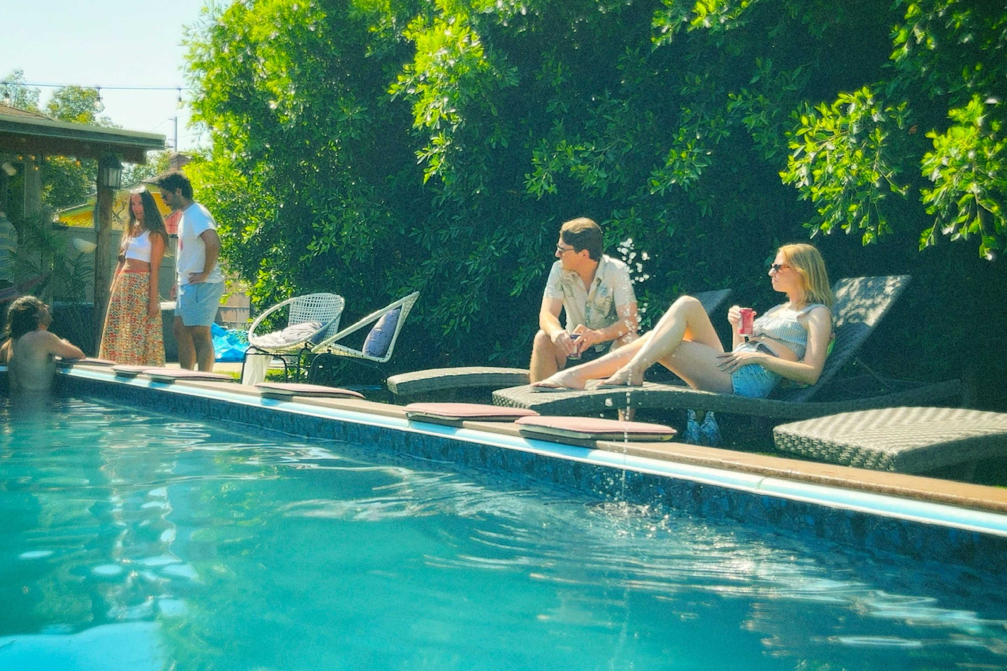 A group of people relaxes by a swimming pool surrounded by greenery, with one couple lounging on poolside chairs.